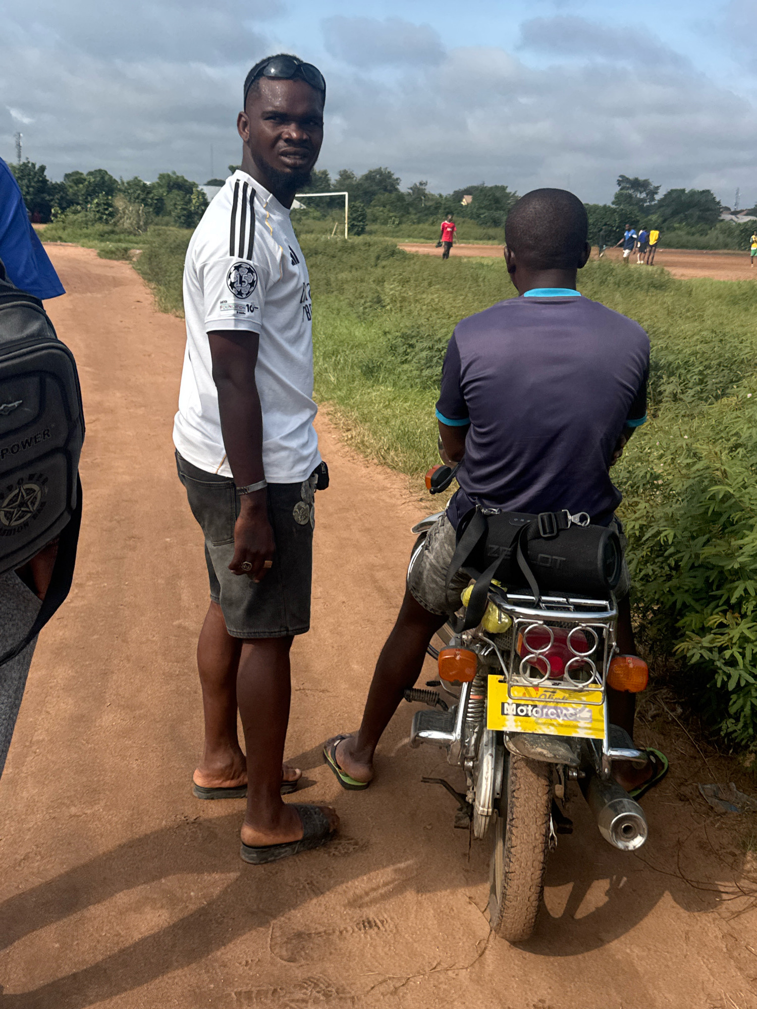 A man wearing a white Adidas shirt and dark shorts stands on a dirt road in Ihiagwa, Nigeria. He has sunglasses on top of his head and is looking at the camera. A boy wearing a blue and purple shirt is sitting on a motorcycle in front of him. The motorcycle has a black bag strapped to the back and a yellow license plate. In the background, there are trees and a soccer field with people playing. The sky is cloudy.