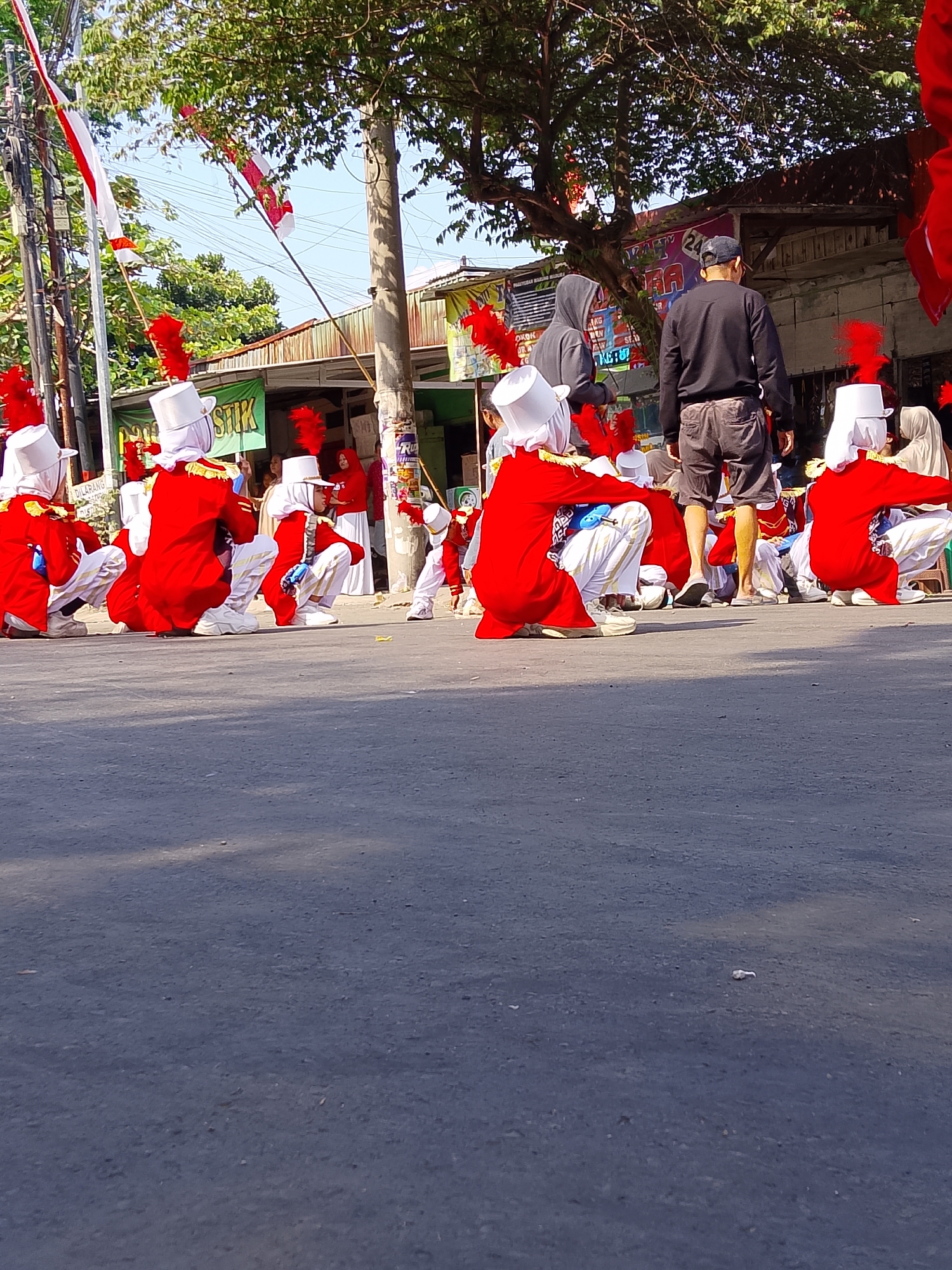 A group of individuals, some in matching red uniforms with white hats and red plumes, are crouched on an asphalt surface. The uniforms appear to be part of a marching band or performance ensemble. Some individuals are wearing headscarves. In the background, there are buildings, trees, and power lines, suggesting an outdoor urban or semi-urban setting. Two adult males are standing in the background, one wearing a black long-sleeved shirt and shorts, and the other a grey hooded sweatshirt. Several red fabric banners or flags are visible, some hanging from poles. The presence of Indonesian flags further suggests the location. The ground is paved and shows signs of wear. The image is taken from a low angle, looking up towards the subjects and the background.