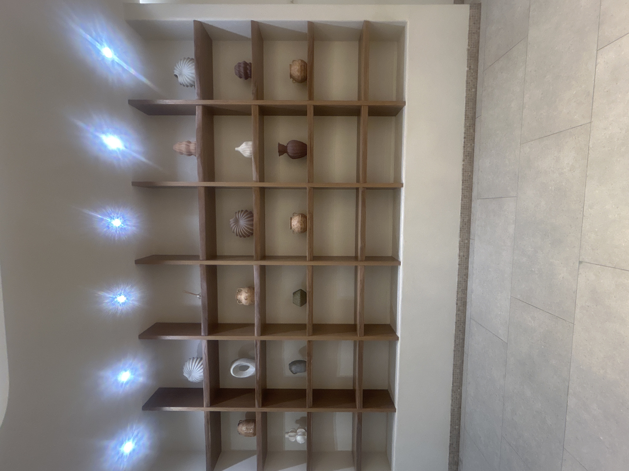 The image shows a well-lit, empty retail display area with multiple shelves. The shelves are made of wood and are arranged in a grid pattern against a light-colored wall. Several decorative items, primarily ceramic vases and sculptures of various shapes and sizes, are sparsely placed on the shelves. The floor is tiled with large, light-colored rectangular tiles. The ceiling features recessed lighting fixtures that cast bright, star-shaped reflections on the ceiling and walls. The overall impression is one of a modern and minimalist design, possibly a section of a showroom or a decorative wall unit in a commercial space. There are no people or visible signs indicating a specific location within Almaty, Kazakhstan, though the design aesthetic is consistent with contemporary interior design trends found in urban environments.