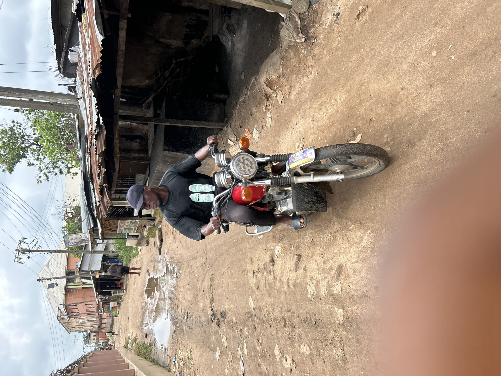 A man is riding a red motorcycle down a dirt road in Ihiagwa, Nigeria. He is wearing a black long-sleeved shirt, black pants, and a blue baseball cap. He is also wearing black sandals. The motorcycle is a red and silver model with a license plate that reads "CZ7237". The road is muddy and there are puddles of water on either side. Buildings line the road, some with corrugated metal roofs. The sky is overcast, suggesting it might be a cloudy or rainy day. The scene appears to be a residential or commercial area with a mixture of permanent and temporary structures. The overall impression is one of a typical day in a Nigerian community, with the motorcycle being a common mode of transportation. There is some debris scattered on the ground, indicating it might be a less-maintained area. The image is oriented vertically, with the man and motorcycle in the center of the frame.