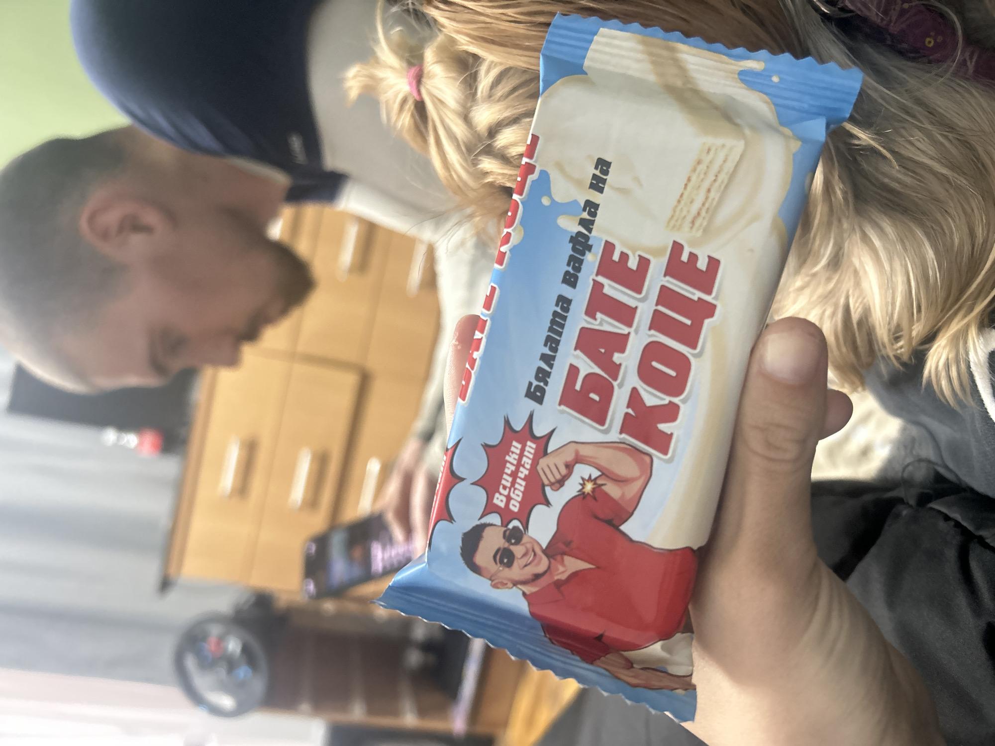 A person holds a "БАТЕ КОЧЕ" wafer bar, a product commonly found in Bulgaria. The packaging is blue and white, featuring an image of a muscular man in sunglasses and a red shirt. The person holding the wafer bar appears to be indoors, possibly at home, with blurred objects like furniture and another person in the background. The specific location in Burgas, Bulgaria, is not discernible from the image.