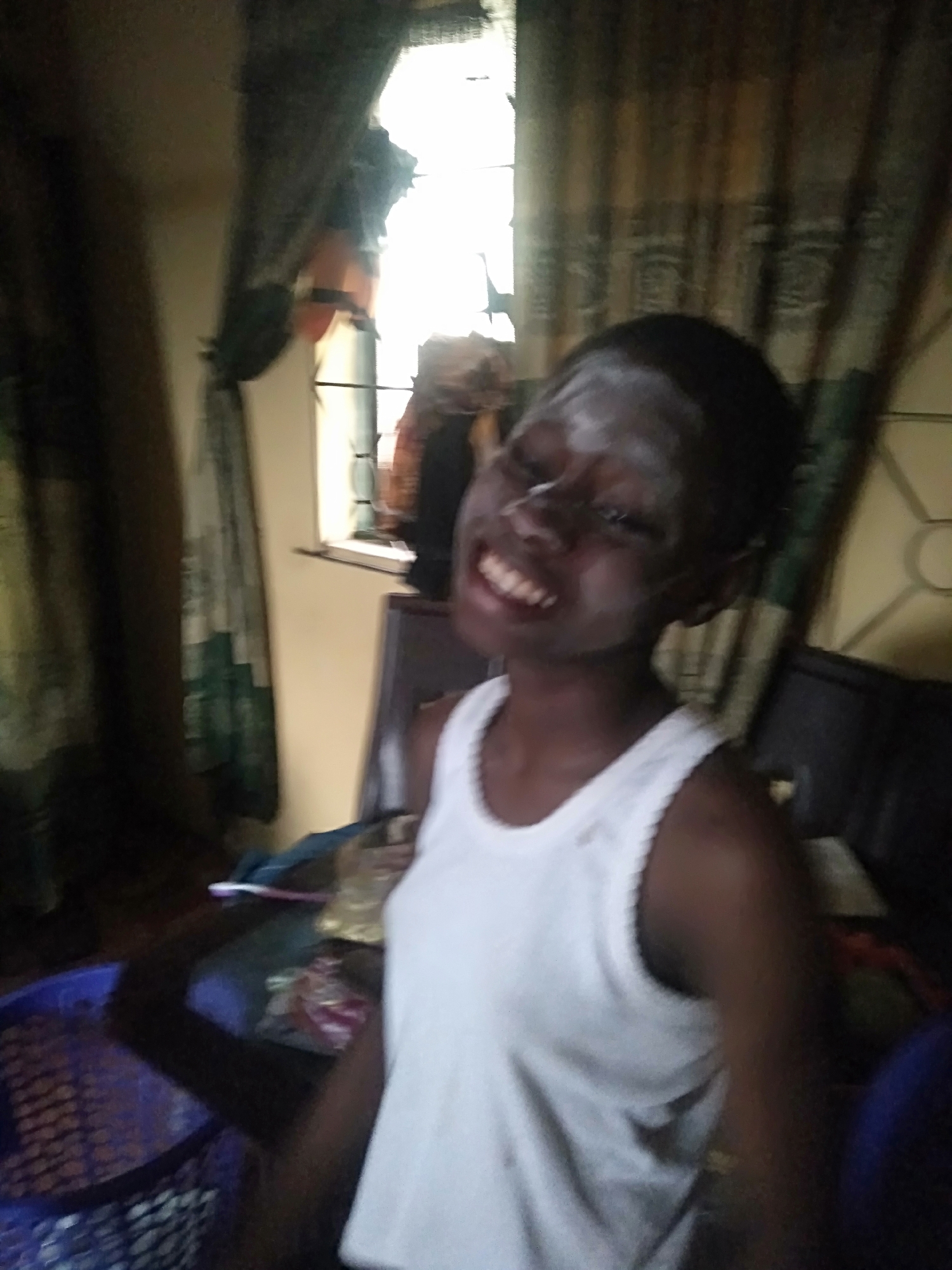 A person with dark skin and white powder on their face is smiling. They are wearing a white tank top. Behind them is a window with patterned curtains. A blue basket is in the lower left corner. The scene appears to be indoors. The location is Shangisha, Nigeria.