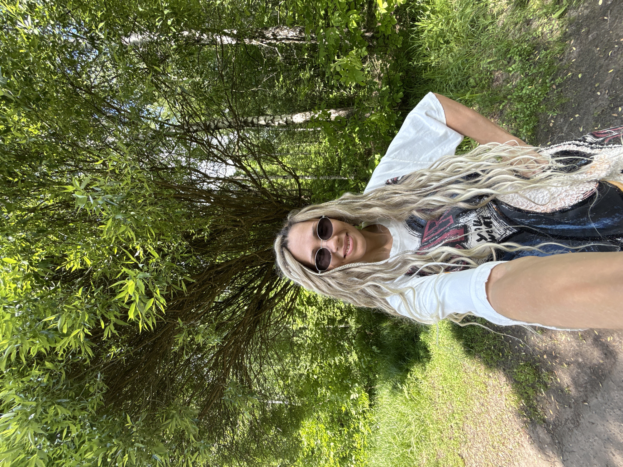 The image captures a vibrant selfie of a young woman in a natural outdoor setting, likely a forest or park in Rusanovo, Russia. The photo is rotated 90 degrees clockwise, showing the woman's head towards the top right of the frame, appearing to be lying on her back relative to the orientation of the trees.
The main subject is a cheerful young woman with long, voluminous blonde hair styled in dreadlocks or thick braids. She is wearing dark, round sunglasses and has a wide, bright smile, indicating happiness. She is dressed in a white long-sleeved t-shirt layered under another dark-colored garment with an abstract or graphic design. Her right arm is extended, holding the camera to take the selfie, while her left hand rests near her hip.
The background is a dense green woodland. A prominent willow tree with numerous thin branches and bright green, slender leaves is very close to the woman on her left side, its foliage creating a rich green backdrop. Further back, more trees with green leaves and visible trunks can be seen. The ground is covered with green grass and a narrow, lighter-colored dirt path or trail.
The photograph was taken during the day under bright sunlight, which casts distinct shadows on the ground and illuminates the scene. The overall impression is one of natural beauty and an enjoyable outdoor experience. There is no clearly legible text visible in the image.