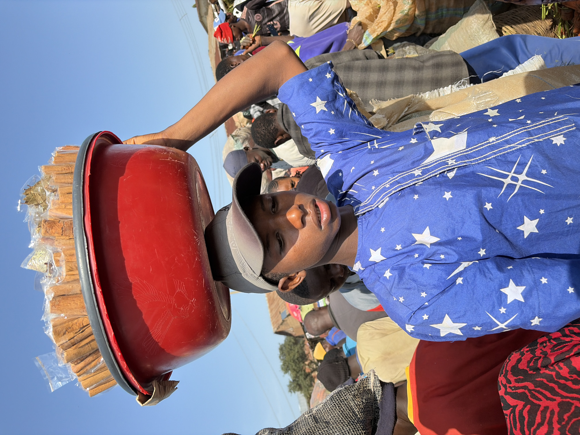 A young boy, likely of West African descent, wearing a blue shirt with white stars and a grey baseball cap, balances a large red plastic basin filled with individually packaged cinnamon sticks on his head. He is walking through a busy outdoor market, possibly in Gura Top, Nigeria, as suggested by the presence of other people and market stalls. The market appears to be bustling with activity, with many people in the background, some carrying goods or engaged in trade. The overall atmosphere seems to be one of commerce and daily life in a Nigerian city. The lighting suggests it is daytime and likely sunny, with a clear blue sky overhead.