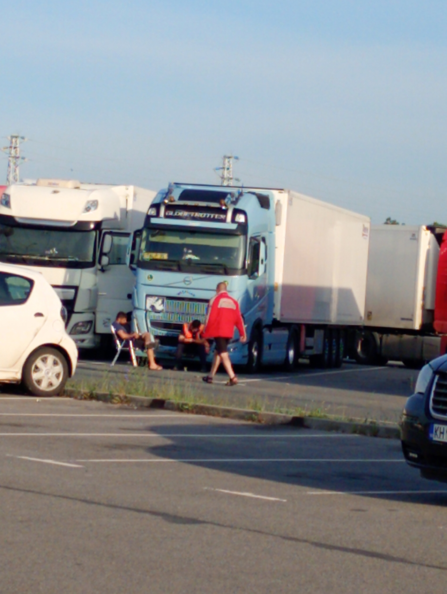 The image depicts a truck stop in Kabile, Bulgaria, during daylight hours. Two semi-trucks, one white and one light blue, are parked adjacent to each other. A white compact car is parked in front of the white truck. A man wearing a red jacket is walking towards the blue truck. Two men are sitting beside the trucks, one in a camping chair and the other on the ground. A black vehicle is partially visible in the lower right corner. Power lines are visible in the background against a clear sky. The ground is paved and shows signs of parking spaces. A strip of vegetation separates the parking area from the trucks.