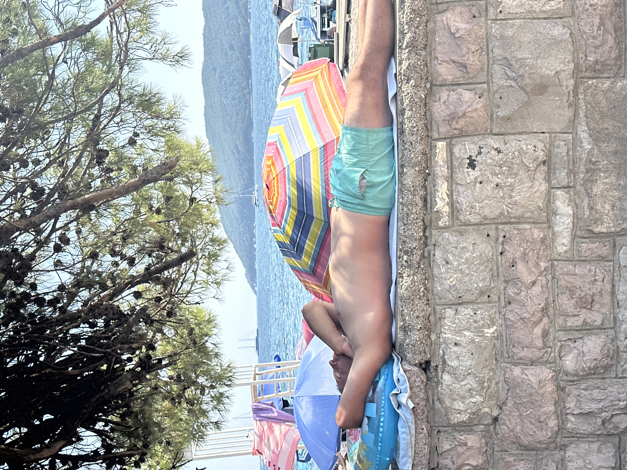 The image captures a sun-drenched scene on a beach, likely in Budva, Montenegro, given the distinctive coastline and mountainous backdrop. In the foreground, a man is lying supine on a stone wall, his head resting on a blue inflatable ring and his body on a white towel. He is wearing light blue-green swim trunks and his eyes are closed, suggesting he is relaxing or sleeping. Behind him, a vibrant, multi-colored striped beach umbrella stands prominently, with a blue umbrella visible to its left. The sea stretches out in the background, dotted with a few sailboats and a distant, hazy mountain range. The upper part of the image is dominated by the dense branches of a pine tree, its needles casting dappled shade over the scene. The stone wall itself is constructed from rough-hewn blocks of varying sizes and colors, adding a rustic texture to the composition. The overall atmosphere is one of peaceful summer leisure.