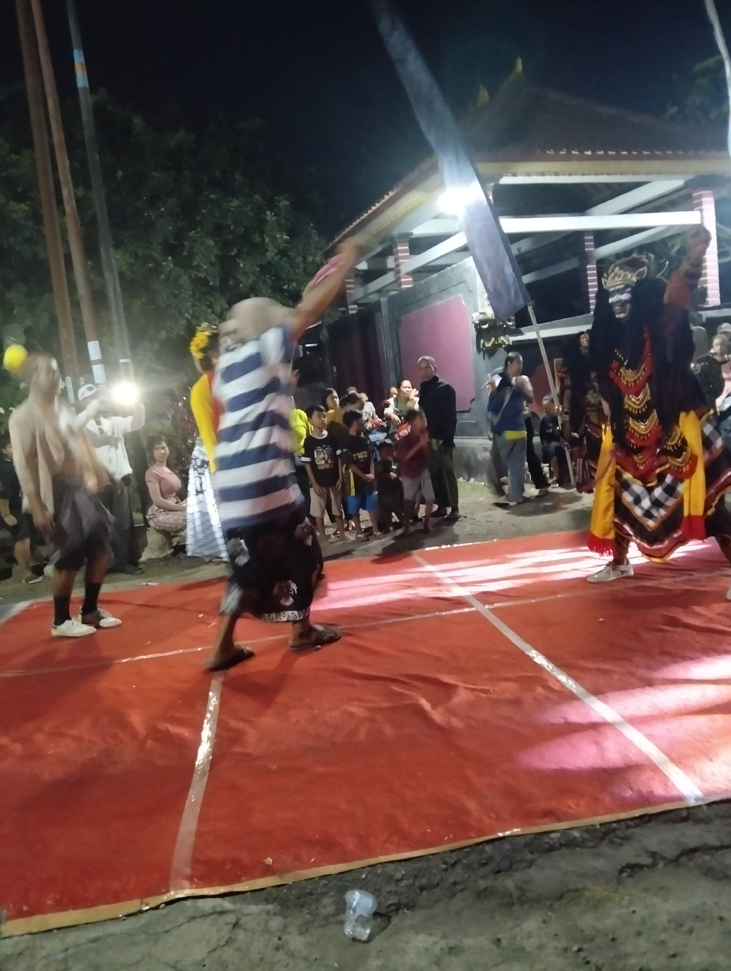 In the village of Sumbersewu, Indonesia, a nighttime cultural performance is taking place outdoors on a raised red stage. The stage is marked with white lines. To the right, a performer in a black and red elaborate costume with a headdress that resembles a demonic face is dancing, holding a black flag attached to a pole. Another performer in the center, wearing a striped shirt and a dark patterned sarong, is also captured mid-motion. A third performer on the left, bare-chested with a dark sarong, is visible. A crowd of spectators, including children and adults, stands behind the stage, illuminated by artificial lighting. The background features trees and a building with a distinctive roofline, indicative of Indonesian architecture. The overall atmosphere is festive and energetic, suggesting a traditional dance or ritual.