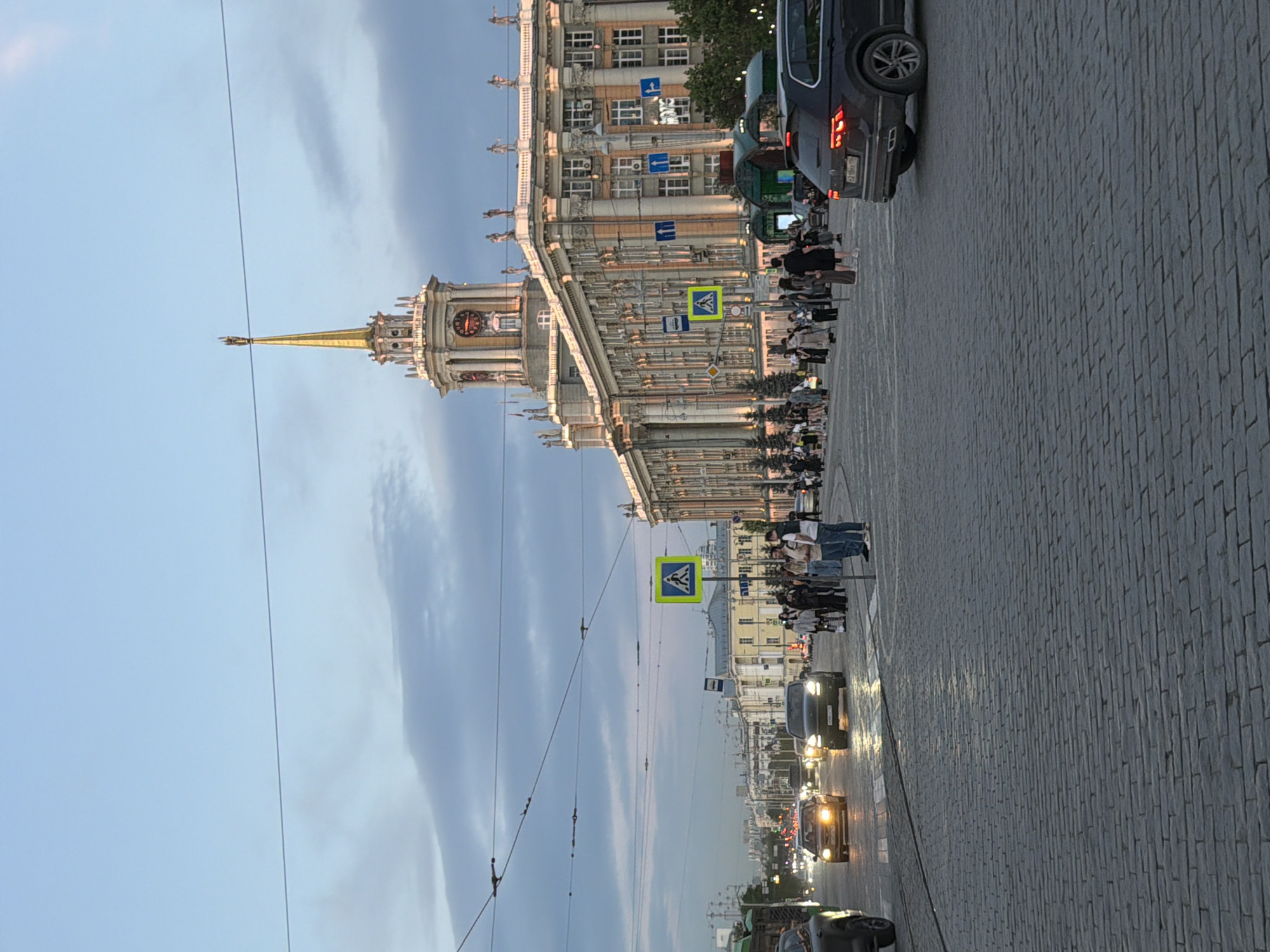The image shows a view of the city of Yekaterinburg, Russia. The main subject is a large, ornate building with a clock tower and golden spire. The building is light-colored and features multiple windows and architectural details. Below the building is a wide pedestrian area paved with cobblestones, where several people are standing and walking. A few cars are visible on the street adjacent to the pedestrian area, including a dark-colored SUV. Pedestrian crossing signs are present along the road. Overhead wires stretch across the sky, which is partly cloudy with a blue hue. The perspective is slightly angled, presenting a view along the length of the street and the building. The scene seems to be lit by the late afternoon or early evening sun.