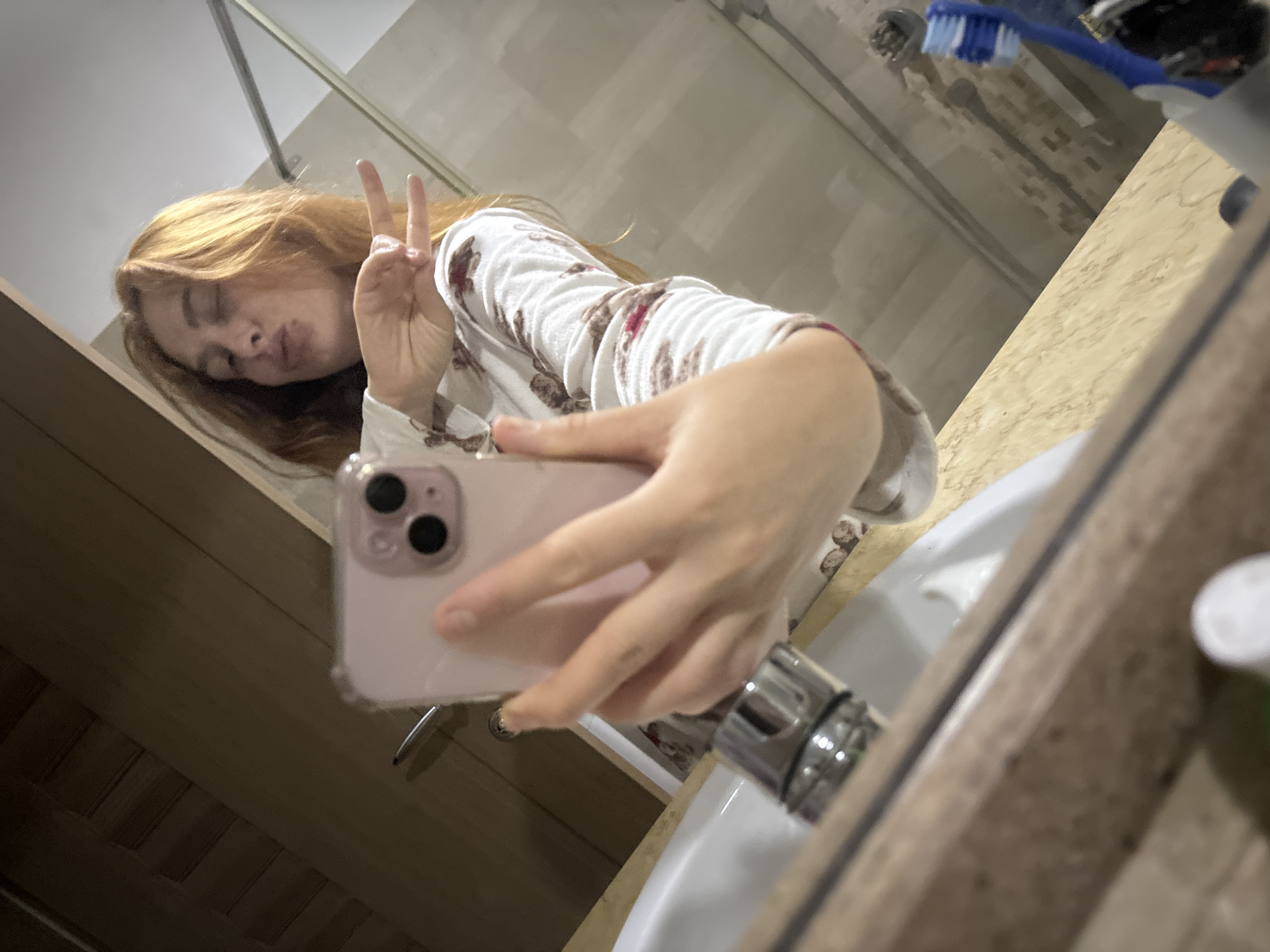 A fair-skinned person with long, auburn hair is taking a selfie in a bathroom. The person is holding a pink smartphone in their right hand. They are making a peace sign with their left hand and pursing their lips in a kissing gesture. They are wearing a white long-sleeved shirt with a brown and red pattern. The bathroom has light-colored walls, a sink with a chrome faucet, and a toothbrush on the counter. The location is likely a private residence or hotel in the city of Budva, Montenegro, based on the general appearance and typical interior design. The setting suggests a routine activity of personal hygiene or self-presentation.