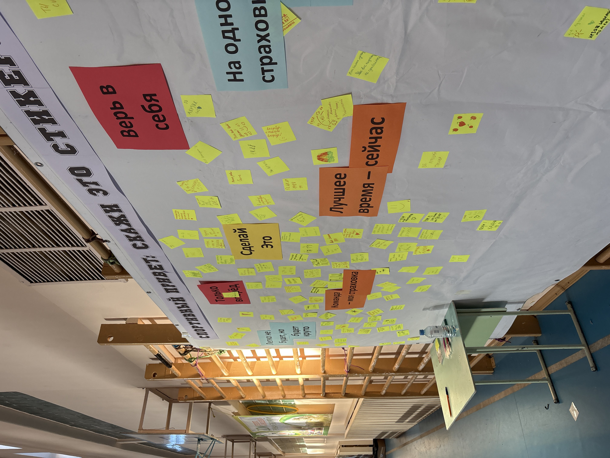The image shows a wall with several colorful posters and numerous yellow sticky notes attached to it. The posters have Russian text, with phrases such as "Believe in yourself," "Only Forward," "Do it," "The best time is now," and "For one insurance." The sticky notes appear to contain handwritten notes or messages, also likely in Russian.
The scene is set indoors, possibly in a school or community center in Berezniki, Russia. There's a glimpse of what seems to be a gymnasium with hanging exercise equipment and a school desk. The lighting appears to be artificial. The composition and content suggest the wall serves as a motivational or interactive display.