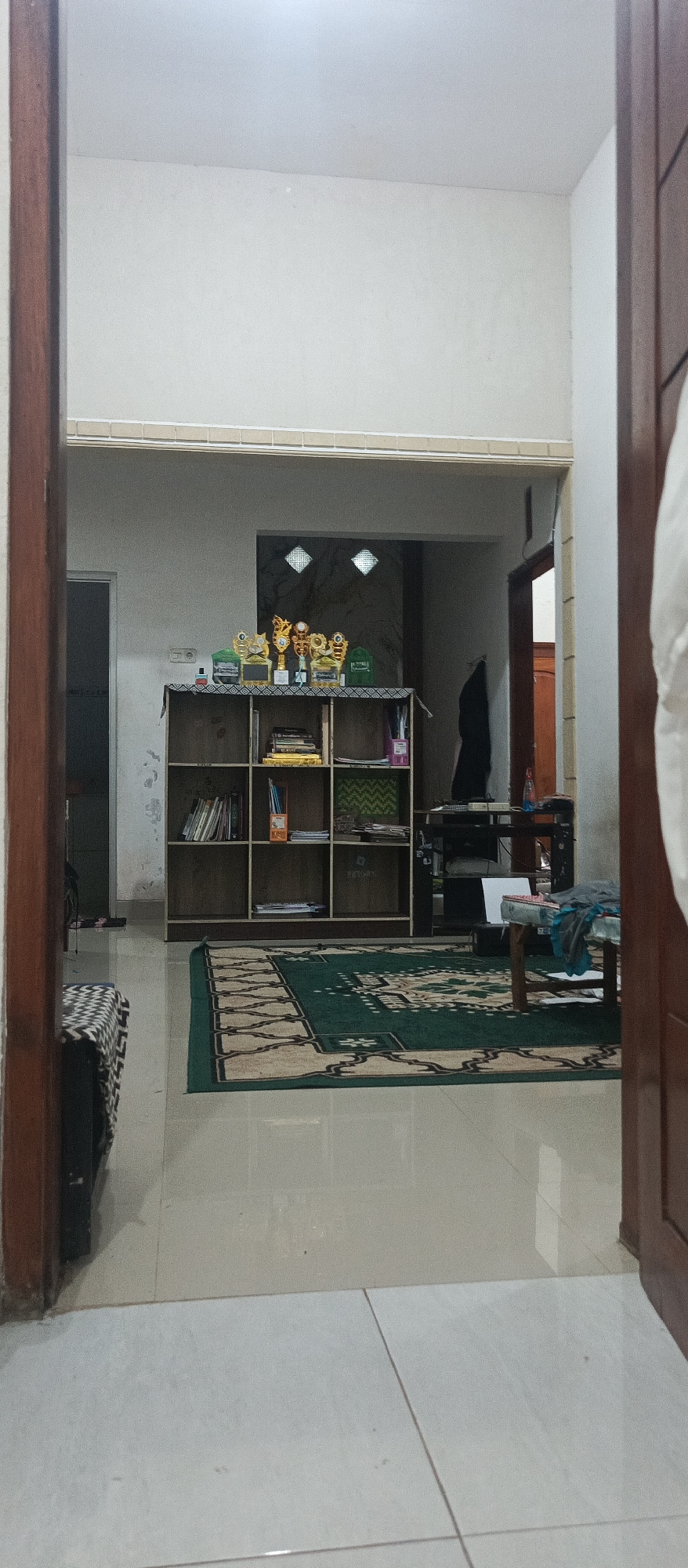 The image shows an indoor scene, possibly a living room or study area. In the center of the frame is a dark wooden bookshelf with multiple compartments. The top shelf displays several trophies and some books. The lower shelves are filled with books and some decorative items. In front of the bookshelf, a green rug with a patterned border covers a portion of the tiled floor. To the left of the bookshelf, a small bench with a patterned cushion is visible. Beyond the bookshelf, a doorway leads to another room, and a black coat hangs on the wall. To the right of the bookshelf, a dark table holds a printer, some papers, and other objects. The overall environment appears to be tidy and well-lit with natural light coming from the side.