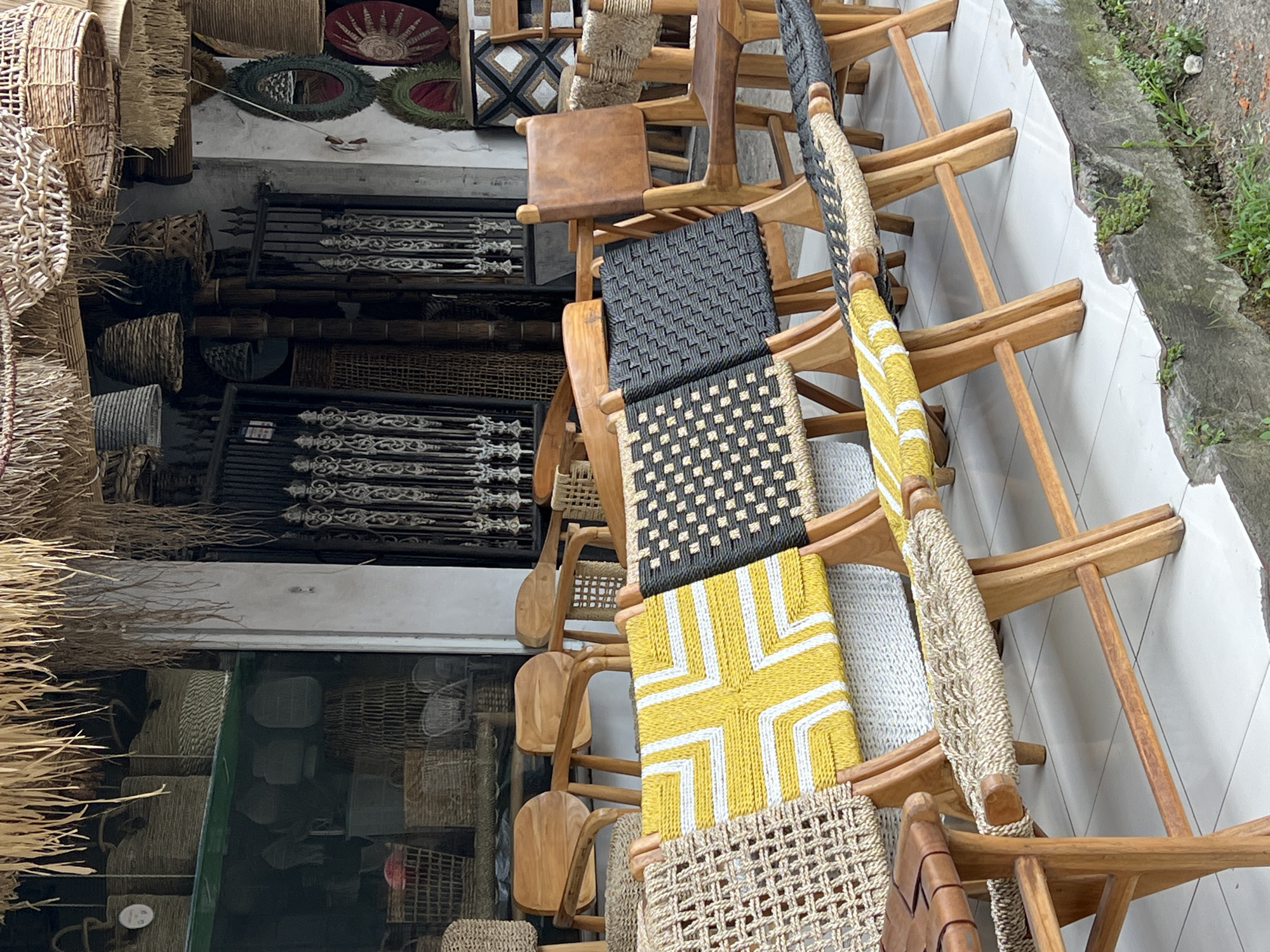 This image captures a vibrant display of handcrafted furniture and decor items, likely within a retail space or workshop in the Ubud District of Indonesia. The scene is dominated by numerous wooden chairs, many featuring seats and backs crafted from various woven natural fibers and materials.
In the foreground, a stack of chairs with light wooden frames showcases diverse seating designs. These include patterns of black and natural-colored woven squares, a prominent yellow and white geometric pattern resembling a cross or plus sign, a light beige woven square pattern, and a solid brown leather seat. Some chair backs are wrapped with natural-colored rope or braided material. To the left, more chairs with carved wooden backs are visible through a doorway.
Beyond the chairs, a dense collection of woven baskets and other decorative items made from natural materials are visible. These include various sized baskets, some hanging and others stacked, and several circular mirrors framed with green and red woven designs. On the wall behind, dark metal objects, possibly decorative railings or gate sections, feature intricate scrollwork.
The chairs are arranged on a clean, white tiled floor. To the far right, beyond the tiled area, a rough concrete edge borders a patch of dirt and small green plants, suggesting an outdoor or semi-outdoor setting adjacent to the display. The lighting indicates it is daytime. No people are visible in the image, and the scene depicts a static display of goods. A small, unreadable circular white tag is affixed to one of the chairs. The abundance of natural materials and handcrafted goods aligns with the artisan culture of Ubud.