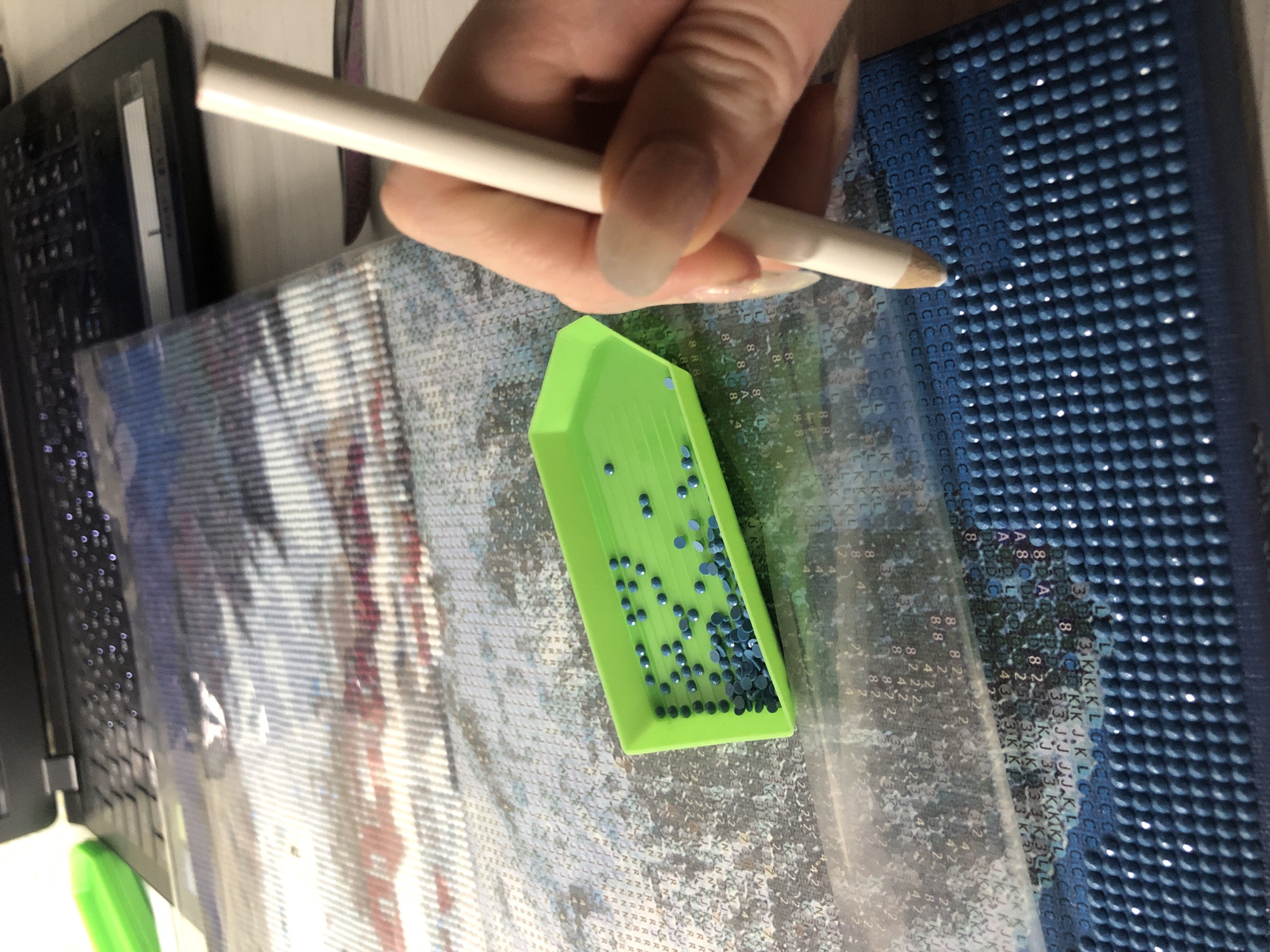 The image shows a person engaged in a diamond painting craft. A hand holds a white stylus above a partially completed canvas. The canvas is covered in a grid of symbols, some of which have been filled with blue diamond beads. A small, green plastic tray holds more of the blue beads. The canvas is placed next to a laptop. A rubber mat is seen on the right side of the image, also featuring tiny diamond beads attached to it.