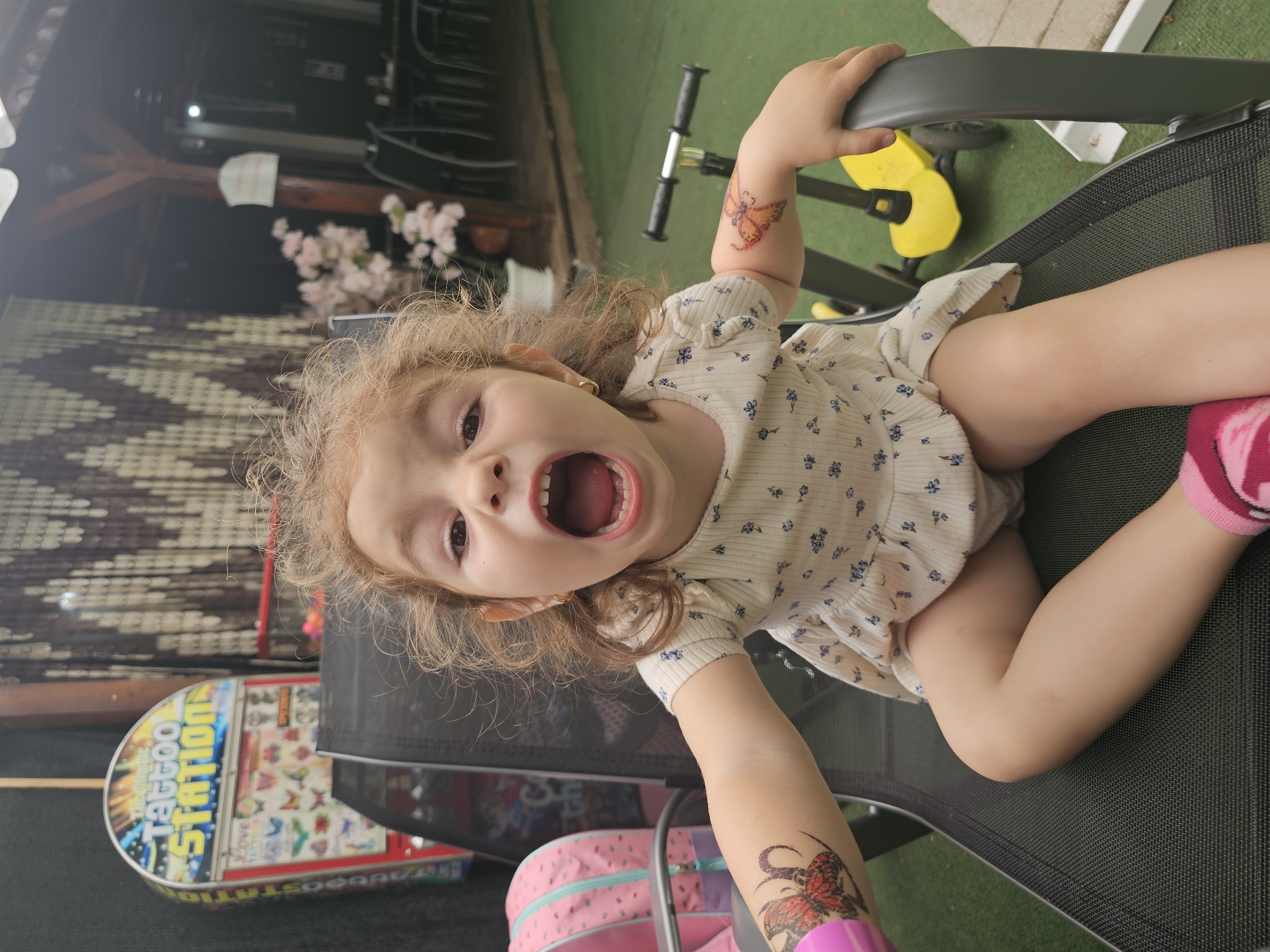 An outdoor scene in Sofia, Bulgaria features a young girl with curly hair sitting in a black mesh chair. She is wearing a light-colored dress with blue floral patterns and pink socks. Temporary butterfly tattoos adorn her arms. Her mouth is wide open in a shout or laugh. In the background, a "Tattoo Station" arcade machine is visible, along with a pink backpack and a yellow scooter. The setting appears to be a backyard or patio area with artificial turf and outdoor furniture.