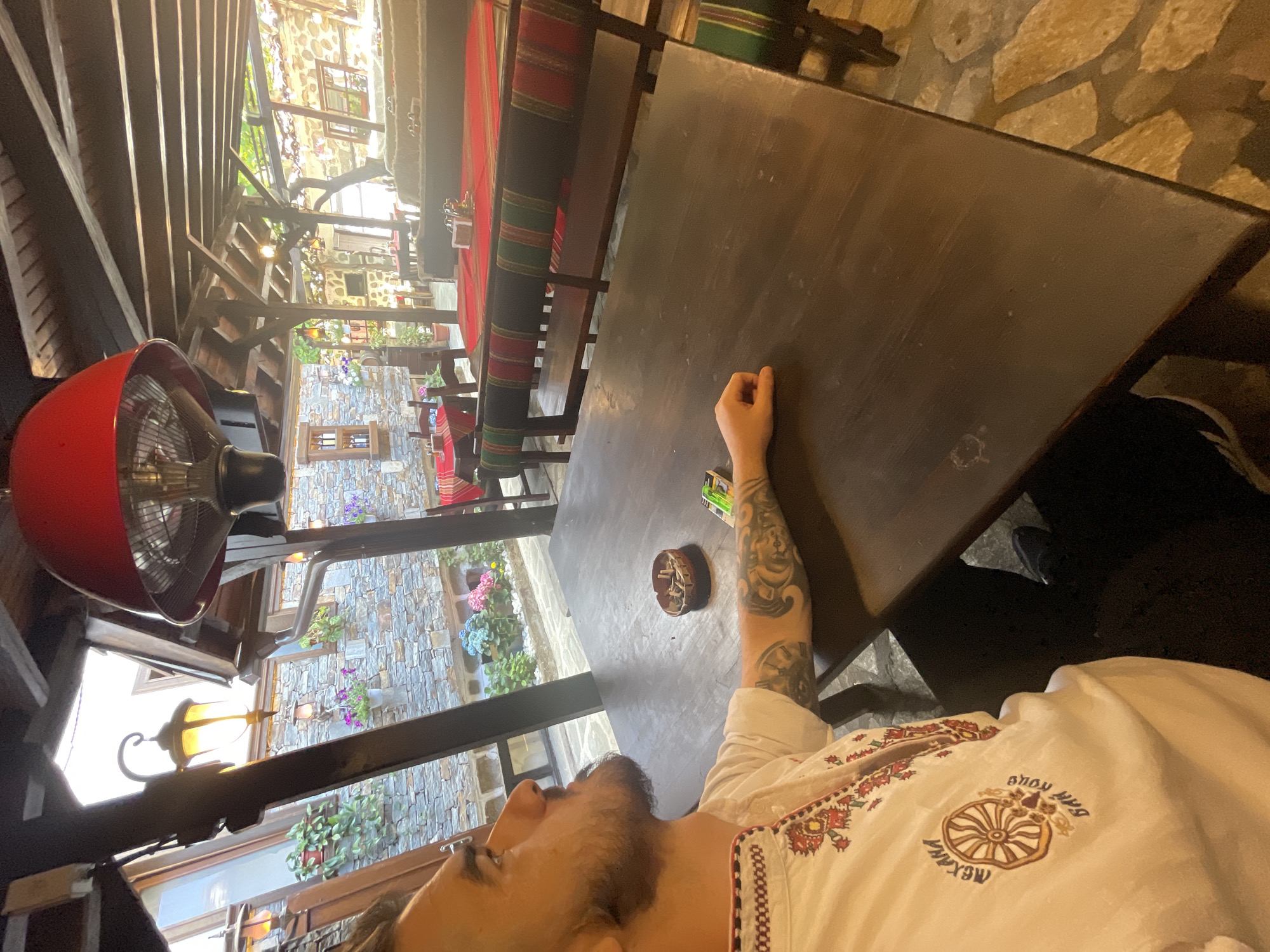 A man with dark hair and a beard sits at a wooden table in an outdoor dining area with a covered roof. He wears a white shirt with intricate embroidered patterns and a circular emblem on the left side. His left arm rests on the table, revealing tattoos. A small ashtray filled with cigarette butts and a pack of cigarettes are on the table in front of him. The background shows a stone building with windows and potted plants, and other tables with red and green striped runners. The lighting suggests it is either early morning or late afternoon.