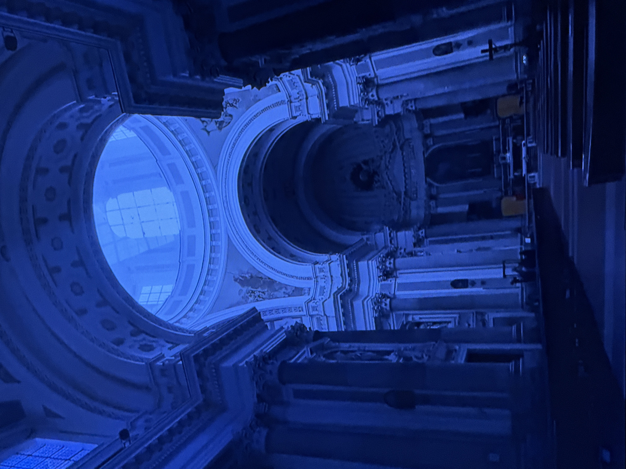 The image shows the interior of a grand, dimly lit church or cathedral, dominated by a deep blue hue, likely due to lighting or a filter. The architecture features a prominent dome with arched windows at the top, elaborate decorative moldings, and tall columns supporting arched ceilings. Rows of pews are visible in the foreground and middle ground, leading towards an altar area. A cross is present on the right side of the altar. The overall impression is one of solemnity and architectural grandeur. There are no discernible people or specific architectural features that would place this scene in Dubai, United Arab Emirates.