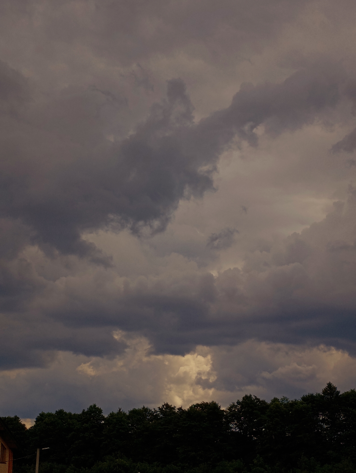 A dark, overcast sky dominates the upper portion of the frame. Heavy, grey clouds are densely packed, with some lighter patches suggesting breaks or thinner cloud cover. The lower portion of the image features a dark silhouette of a dense line of trees, forming a natural horizon. A sliver of a building, possibly a house with a brick facade, is visible on the far left. A thin, vertical pole, likely a lamppost, stands just to the right of the building. The overall impression is of an impending storm or the aftermath of one. The image does not contain any people or discernible actions. There are no specific landmarks visible that would definitively place this scene in Minsk, Belarus.
