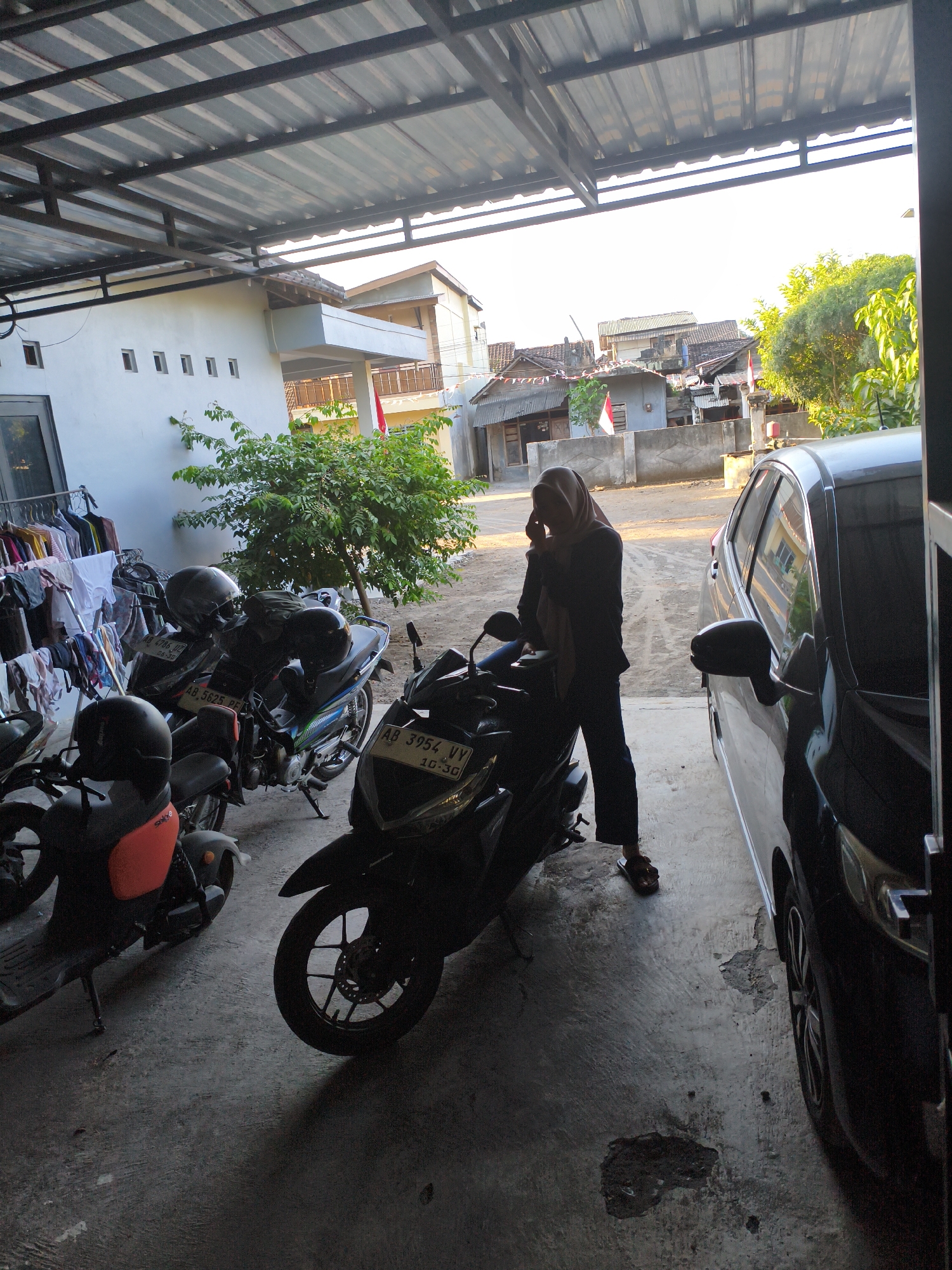 The image depicts an outdoor scene in Berbah, Indonesia, likely a residential area or a parking space. A covered area, possibly a carport, is visible with a corrugated metal roof supported by metal beams. In the foreground, several motorcycles are parked. One motorcycle, a black scooter, has a license plate reading "AA 3954 VV". Another motorcycle, a dark-colored scooter with an orange rear section, is also present. To the right, a black car is parked.
A woman wearing a light brown hijab and dark clothing is standing between the motorcycles and the car, holding her phone to her ear. She is wearing sandals. Laundry is hanging on a rack to the left of the scene.
In the background, houses with tiled roofs and walls of various colors are visible. A tree with green foliage is also present. The ground is paved with concrete. The lighting suggests it is either early morning or late afternoon, with sunlight casting shadows.
