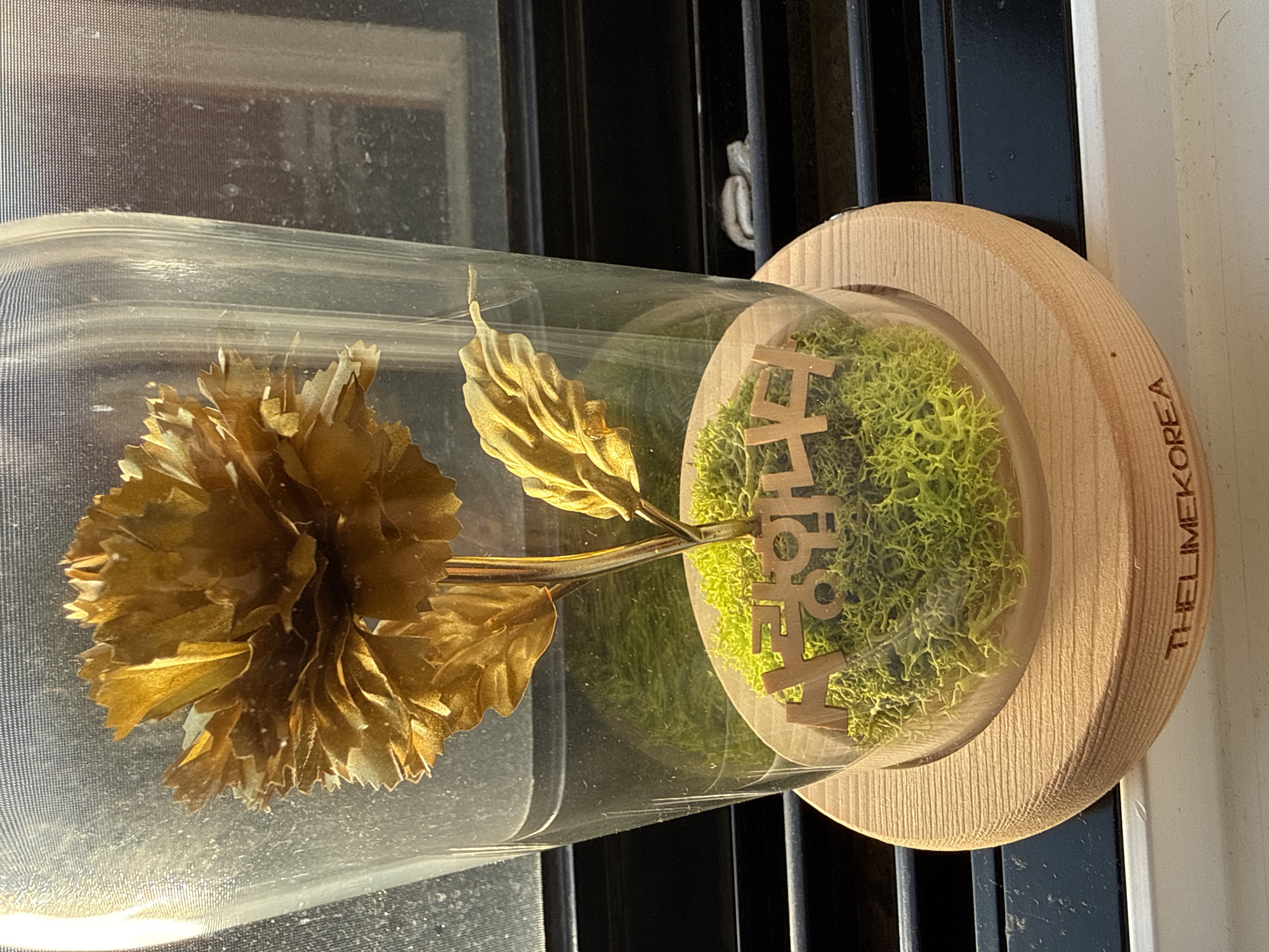 A golden flower under a glass dome sits on a wooden base. The flower has a metallic sheen and is surrounded by green moss. The base has the text "THELIMEKOREA" engraved on it. The background shows a window with blinds, suggesting an indoor setting. There is no indication of the city of Seoul or any specific location within it.