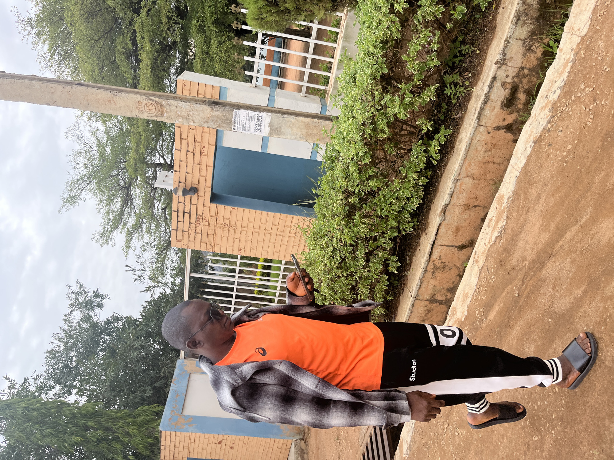 A man is standing outdoors, looking at a mobile phone in his hand. He is wearing an orange t-shirt, black and white pants with the word "Studios" printed on the leg, a gray plaid shirt, dark-rimmed glasses, and black sandals. He appears to be on a concrete surface near a low wall with green bushes growing behind it. In the background, there is a brick structure with a blue door and a white fence. There are trees visible, suggesting an outdoor, possibly urban, location. The image appears to be taken during the day under overcast skies. Given the prompt's requirement for the location in Jos, Nigeria, the scene would be consistent with an urban environment in Jos, Nigeria.