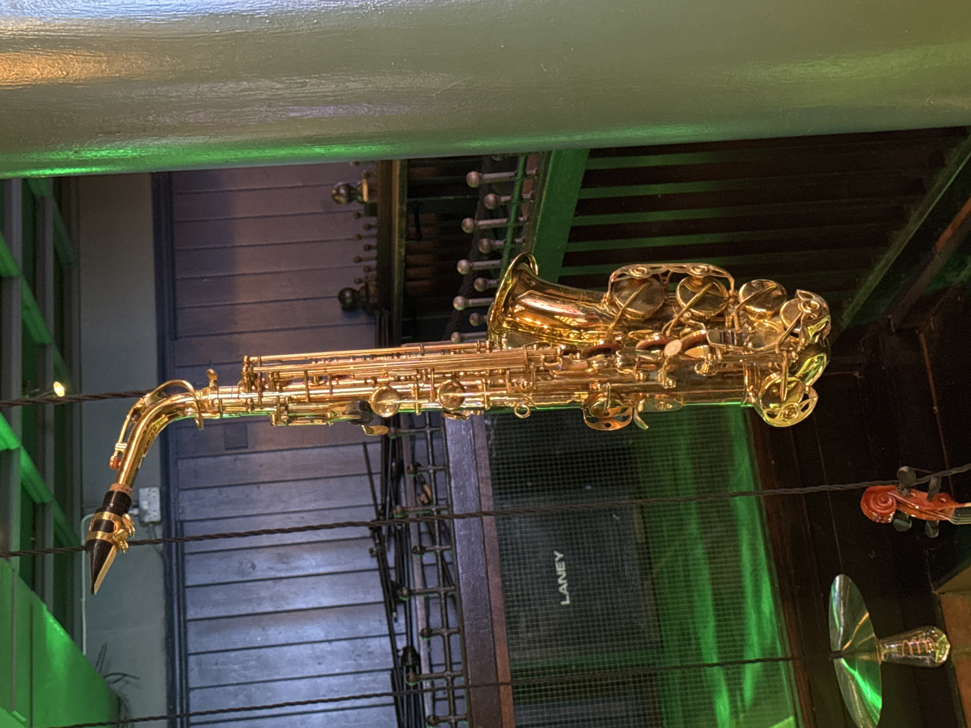 A golden saxophone is prominently displayed, suspended horizontally in the center of an indoor scene, likely a music-themed venue or bar. Its brass surface gleams, reflecting the surrounding light. The saxophone's mouthpiece points to the left, and its bell section, rich with intricate keys and mechanisms, is towards the right.
In the background, dark wooden panels or slats are visible, suggesting a rustic or industrial aesthetic. Below the saxophone, towards the right, a black speaker or amplifier is partially visible, bearing the white text "LANEY" vertically on its grille. Further to the right, a small, dark red scroll and tuning pegs of a string instrument, possibly a violin or cello, are suspended, and below it, a reflective, cymbal-like object or decorative light fixture can be seen.
The scene is dimly lit overall, with a strong artificial green light emanating from the left, casting a vibrant glow on a light-colored beam running across the top of the frame and illuminating the area directly below the instruments. Several dark wires or ropes crisscross the space, appearing to suspend the instruments or act as decorative elements. No people are visible. The atmosphere suggests evening or nighttime, characteristic of entertainment venues.