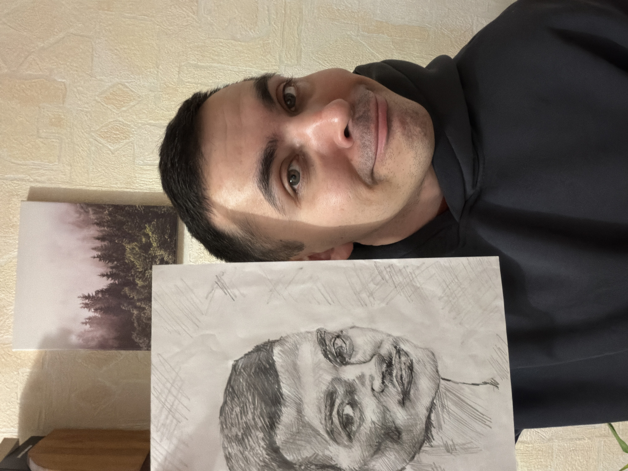 A man wearing a dark hoodie is holding a drawing of a face. The drawing is in black and white and appears to be a sketch. A picture of a forest scene hangs on the wall behind him to the left. The background wall appears to be textured. There is no evidence in the image that explicitly indicates the location is Izhevsk, Russia.