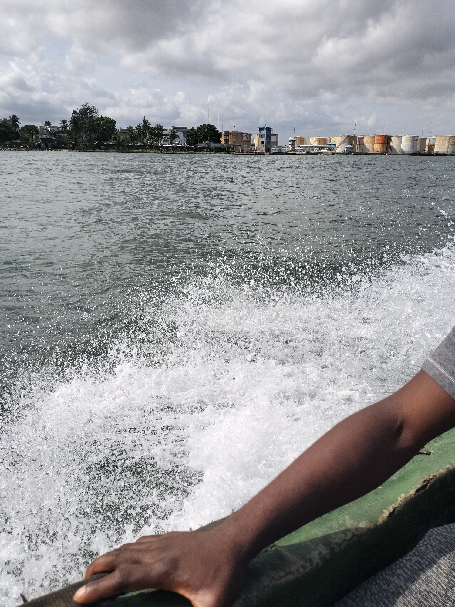 This image captures a view from a moving boat on a body of water, likely a lagoon or river, under an overcast sky. In the foreground, the right arm and hand of a dark-skinned person are visible, resting on the green edge of the boat. The boat's motion creates a prominent white wake and spray that churns the dark grey-green water in the lower and central parts of the frame.
The distant shoreline reveals a mix of development. To the left, a cluster of green trees, including palm trees, partially conceals buildings. To the right, the landscape transitions to an industrial area featuring a blue-roofed structure and a long line of large, cylindrical storage tanks. These tanks display various shades of yellow, orange, and white/grey, suggesting they are used for storing oil or other industrial liquids, indicative of a port or refinery facility.
The sky is covered with dense, grey clouds, indicating an overcast daytime. The scene depicts an active waterway in an urbanized coastal or riverine environment. While text might be present on the distant tanks, it is not clearly legible. The overall setting is consistent with a location such as Lagos, Nigeria, known for its busy waterways and port activities.