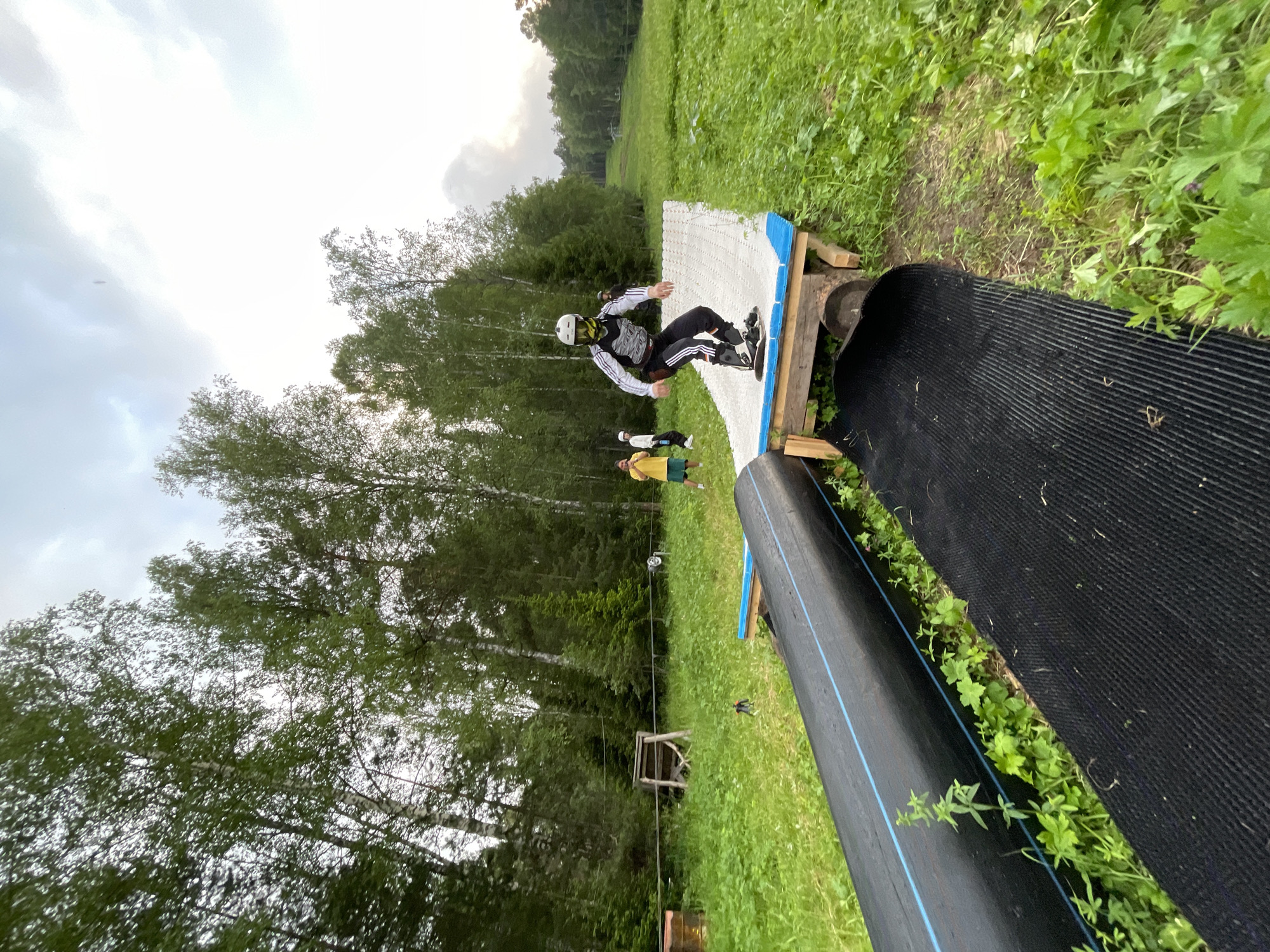 An individual wearing a helmet, goggles, and protective gear is shown on a makeshift ramp constructed from wood and a textured mat. The person is on a snowboard, appearing to be in motion. Behind them, two other individuals stand on a grassy slope, one wearing a yellow shirt and the other in white. The scene is set outdoors, surrounded by lush green grass and tall trees, suggesting a natural environment. In the background, a ski lift or similar structure is visible on a distant hill, indicating the location might be a ski resort or a recreational area. The sky is overcast with clouds. The overall impression is of an outdoor activity, possibly training or practice, taking place in a summer or early autumn season. This image depicts a recreational area in Divnogorsk, Russia, potentially a ski resort or a park, given the presence of a ski lift and the mountainous terrain.