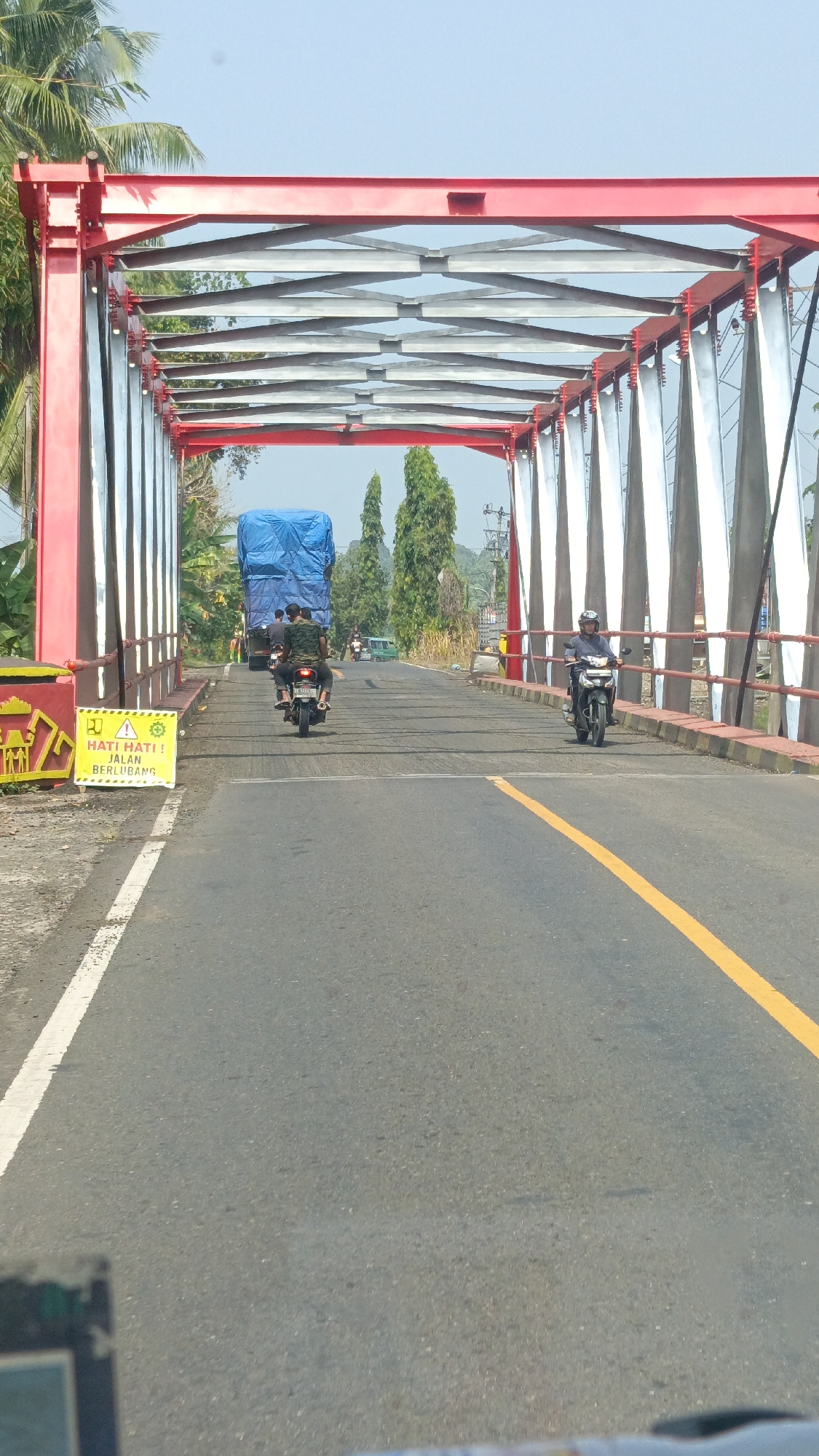 The image shows a view from a vehicle on a road approaching a red truss bridge in Way Gelang, Indonesia. The bridge has a red metal frame with silver-colored horizontal beams and vertical supports. On the left side of the road, just before the bridge, is a yellow and green sign with text in Indonesian that translates to "BE CAREFUL! THE ROAD IS POTHOLED." Two motorcycles are traveling on the bridge. The first motorcycle, closer to the camera and on the left side of the lane, carries two people. The second motorcycle, further ahead and on the right side of the lane, is ridden by a single person wearing a helmet. In the background, a large blue-covered truck is visible, along with other vehicles and trees. The sky is clear and blue.