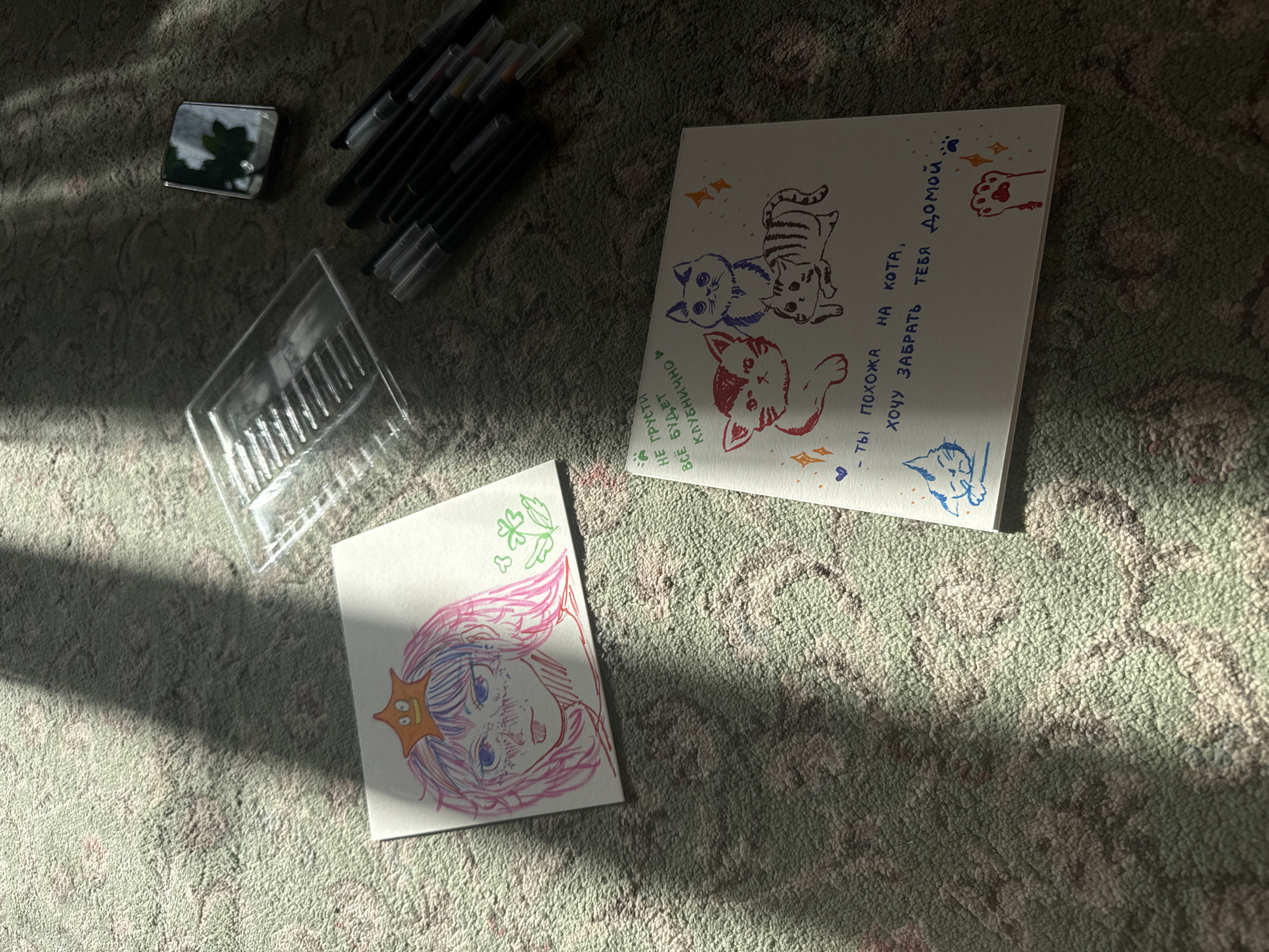 The image captures an indoor scene in Uryupinsk, Russia, with direct sunlight casting sharp shadows across a light green and gray patterned carpet. The angle of the sunbeams suggests either morning or late afternoon.
The primary subjects are two pieces of paper displaying vibrant drawings. The larger drawing features three stylized cats: one blue, one red, and one striped brown, alongside various decorative elements like hearts, stars, and paw prints. Handwritten Russian text accompanies the illustrations. On the left, it reads, "НЕ ГРУСТИ ВСЁ БУДЕТ КЛУБНИЧНО" (Don't be sad, everything will be strawberry). On the right, it says, "- ТЫ ПОХОЖА НА КОТА, ХОЧУ ЗАБРАТЬ ТЕБЯ ДОМОЙ." (- You are like a cat, I want to take you home.).
The smaller drawing, situated to the lower left, depicts an anime-style character with flowing pink and blue hair and large blue eyes, seemingly smiling. A small yellow star sticker is affixed to the character's hair, and faint green clover or leaf designs are drawn in the top corner.
Around the drawings are art supplies: a collection of black markers or pens with colored caps, and a clear plastic container, possibly for holding pens. In the upper left corner, a small, silver-framed mirrored object reflects a blurry image of green foliage. The scene evokes a feeling of creative leisure and conveys comforting and affectionate sentiments through the illustrations and Russian text.