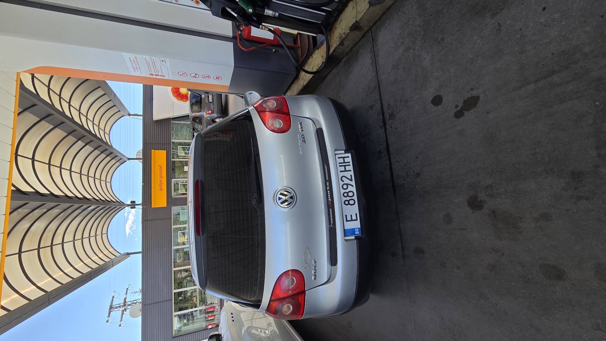 A silver Volkswagen Golf is parked at a gas station, facing away from the camera. The car has a Bulgarian license plate that reads "E 8892 HH". The car is parked under a large, arched canopy made of translucent material supported by metal beams. To the right of the car is a gas pump with several hoses and a fire extinguisher. In the background, a store or building with large windows is visible, and a sign with "добре дошли!" (Welcome!) in orange lettering is mounted above the entrance. The ground is paved with concrete, and there are some dark stains on the ground. The sky is blue with some clouds.