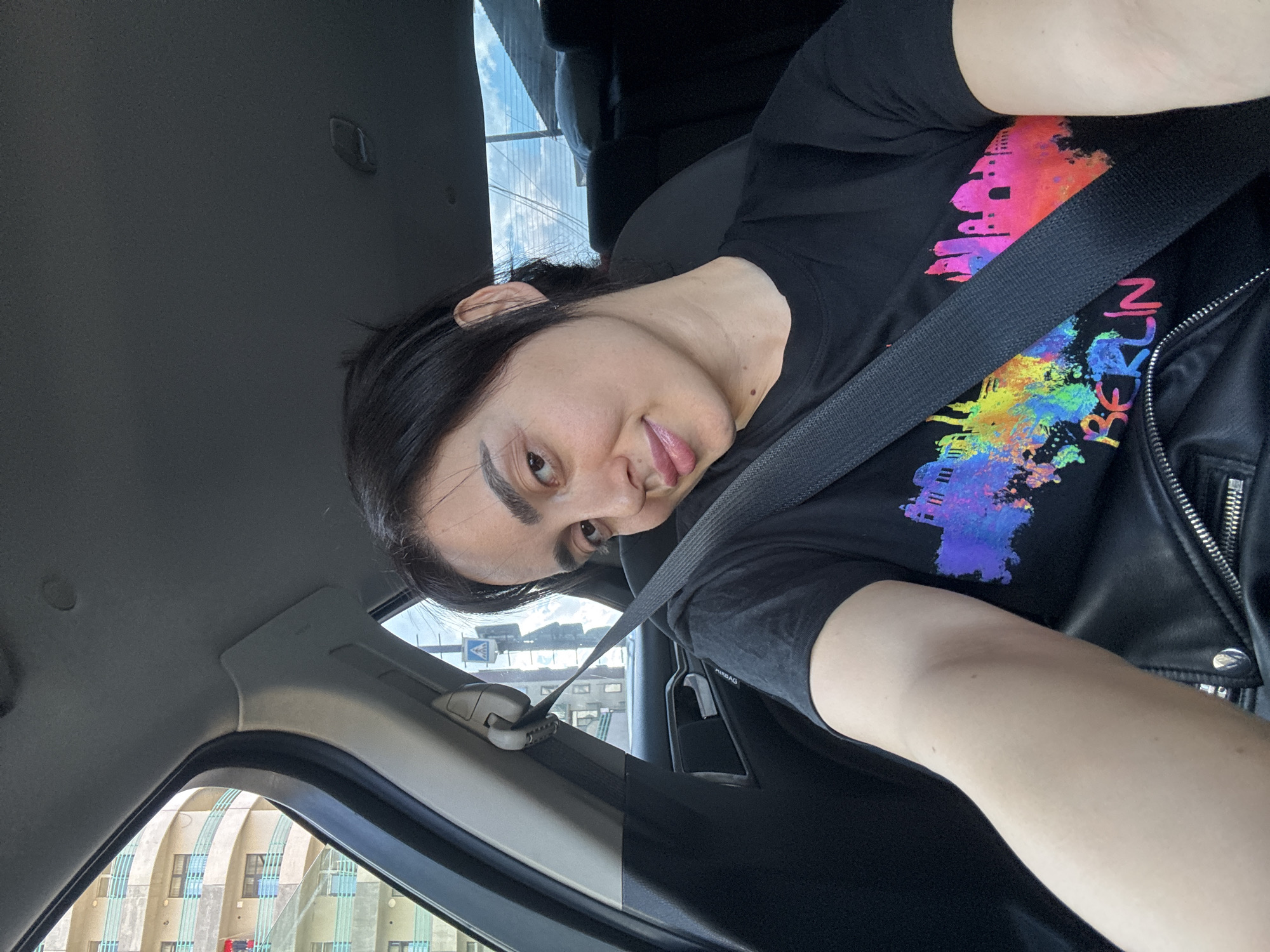 A passenger in a car wearing a Berlin-themed shirt and black leather jacket, with a seatbelt across the chest and shoulder, looking at the camera. The car's interior is visible, with the window showing an exterior scene. Outside the car is a building with arched architectural elements, and road signage is also visible. The scene appears to be in Tbilisi, Georgia.
