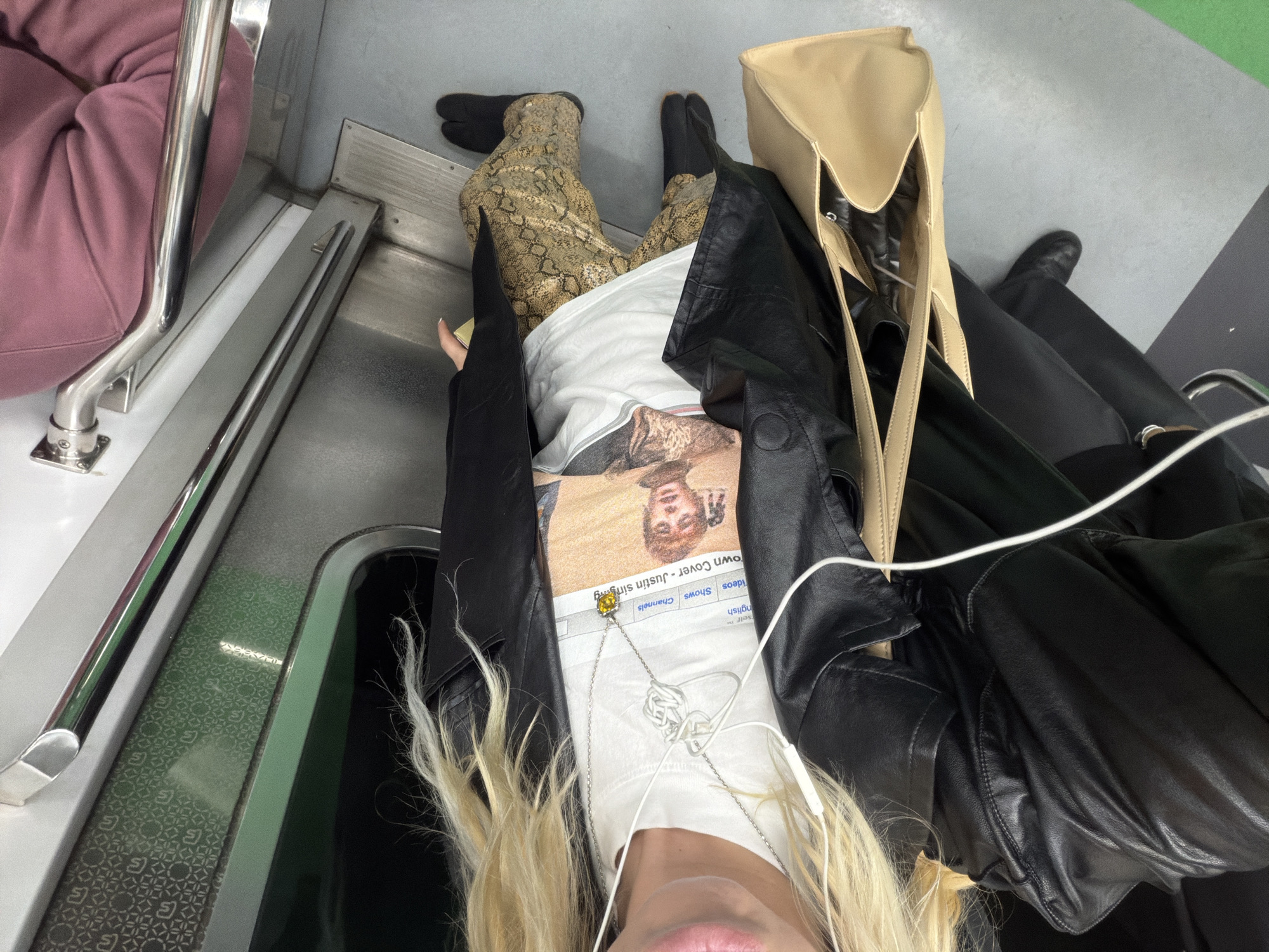 The image depicts a person seated on a train in Seoul, South Korea. They are wearing a white t-shirt with a graphic of Justin Bieber, a black leather jacket, tan snake print pants, and black split-toe shoes. A tan tote bag is next to them. The train interior features a silver handrail, patterned green paneling, and a gray floor. Another person is seated next to them, wearing a pink top.