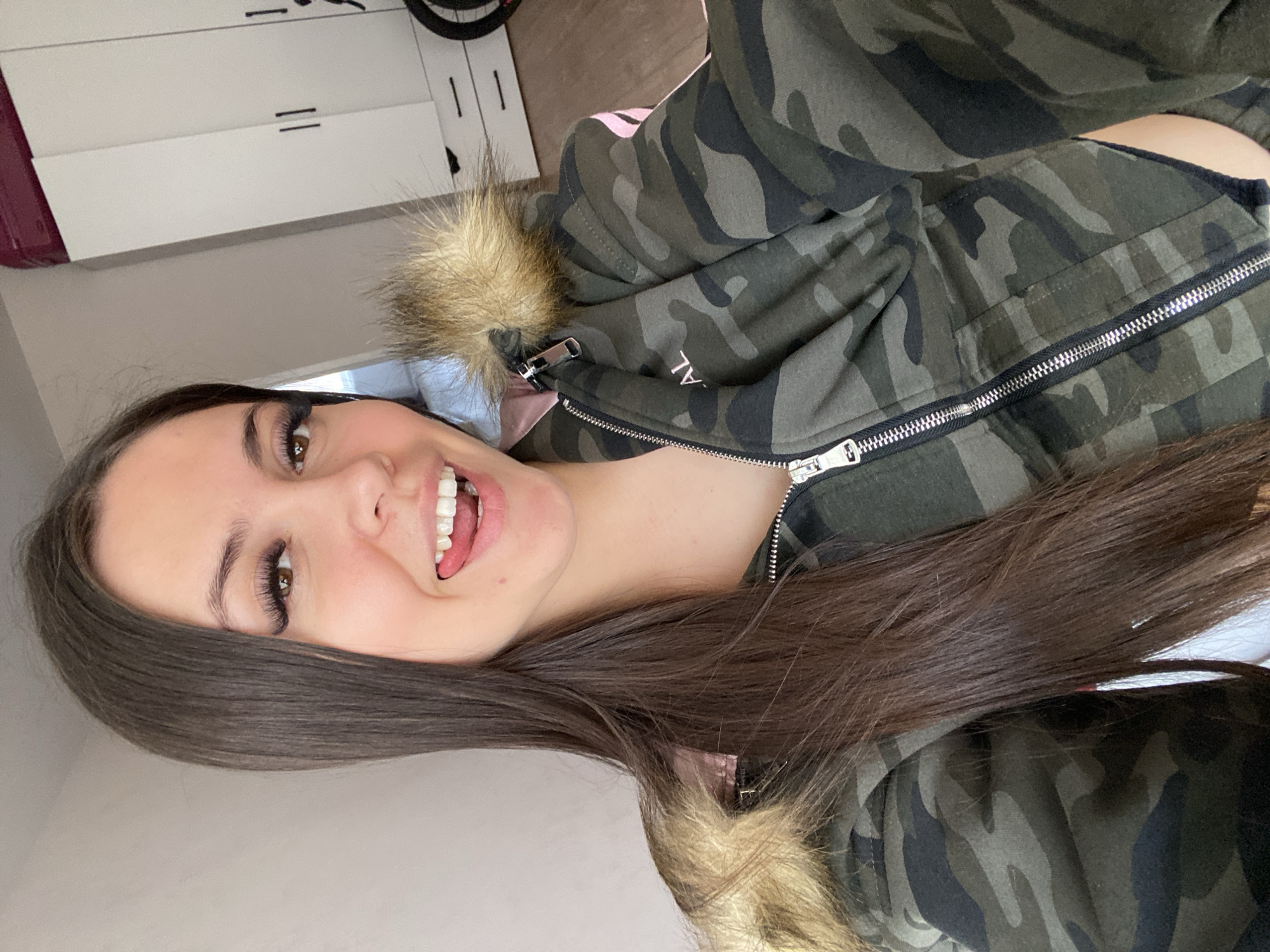 A close-up portrait features a young woman smiling broadly at the camera, with her mouth open revealing her teeth and tongue, suggesting a cheerful or playful mood. Her long, straight dark brown hair frames her face and extends over her left shoulder. She has brown eyes with noticeable eyelashes, possibly extensions.
She is wearing a dark green camouflage-patterned jacket or hoodie, which is partially zipped, revealing a glimpse of a light pink garment underneath. The jacket has a prominent silver zipper and a yellowish-brown fur trim around the collar or hood. On the upper right side of her jacket, part of a word, "AL," is visible.
The setting appears to be indoors, likely a room, with ambient daylight. The background on the left shows white cabinets or drawers with dark horizontal handles. A light-colored wall forms the rest of the backdrop, and a dark, circular object, possibly a bicycle wheel, is partially visible in the upper right. This image is associated with Chaikovsky, Russia.