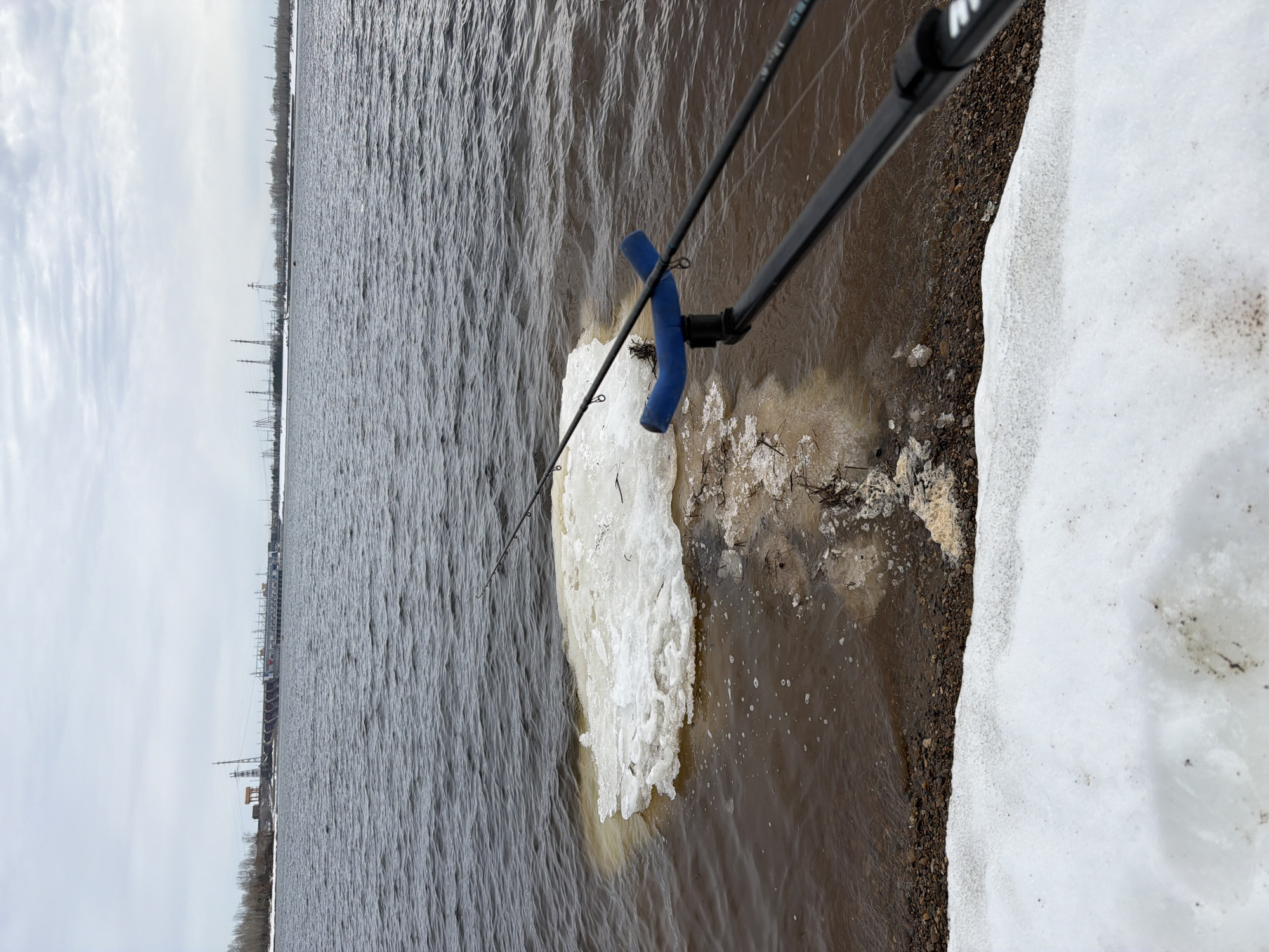 The image depicts a fishing rod in a river in Chaikovsky, Russia. The fishing rod is black and has a blue foam grip. It is positioned near the shoreline where there is snow and ice. The river is murky and choppy. In the background, there are structures including electrical towers, and a grey sky is visible.