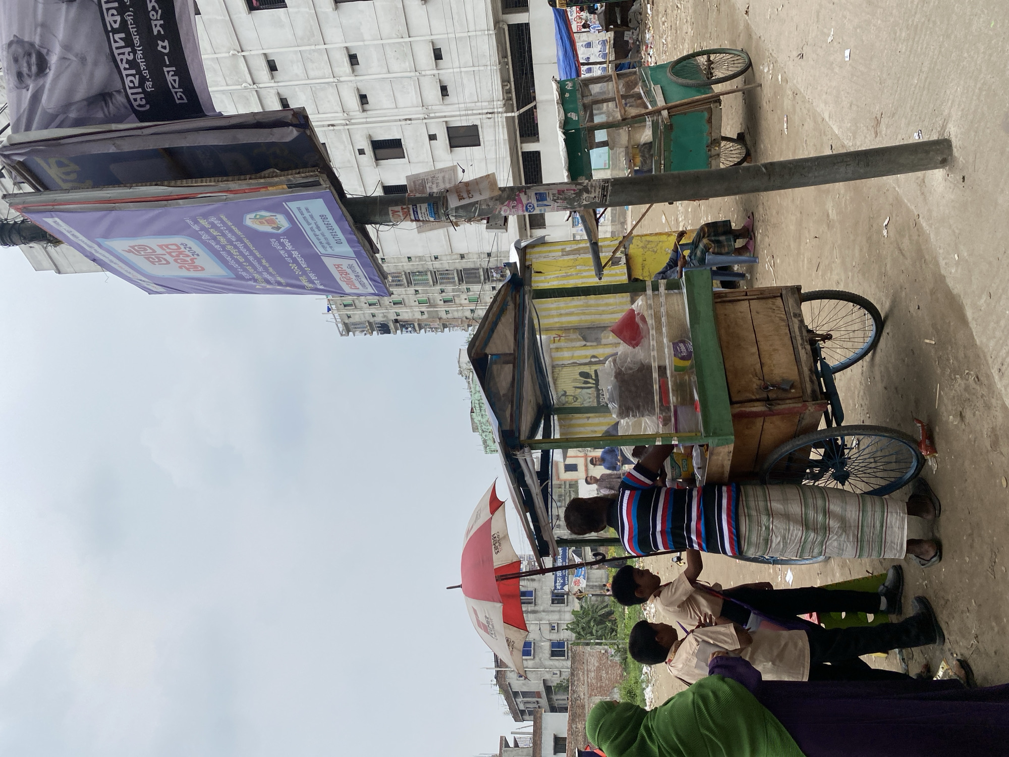 The image depicts a street scene in Dhaka, Bangladesh. A vendor wearing a striped shirt and a lungi stands behind a small cart, possibly selling food or drinks. Two children in school uniforms stand at the cart as well. An umbrella is open next to the cart. Additional carts are on the right side of the image. The background shows buildings, some appearing to be residential or commercial. A light pole stands in front of the vendors with promotional banners attached.