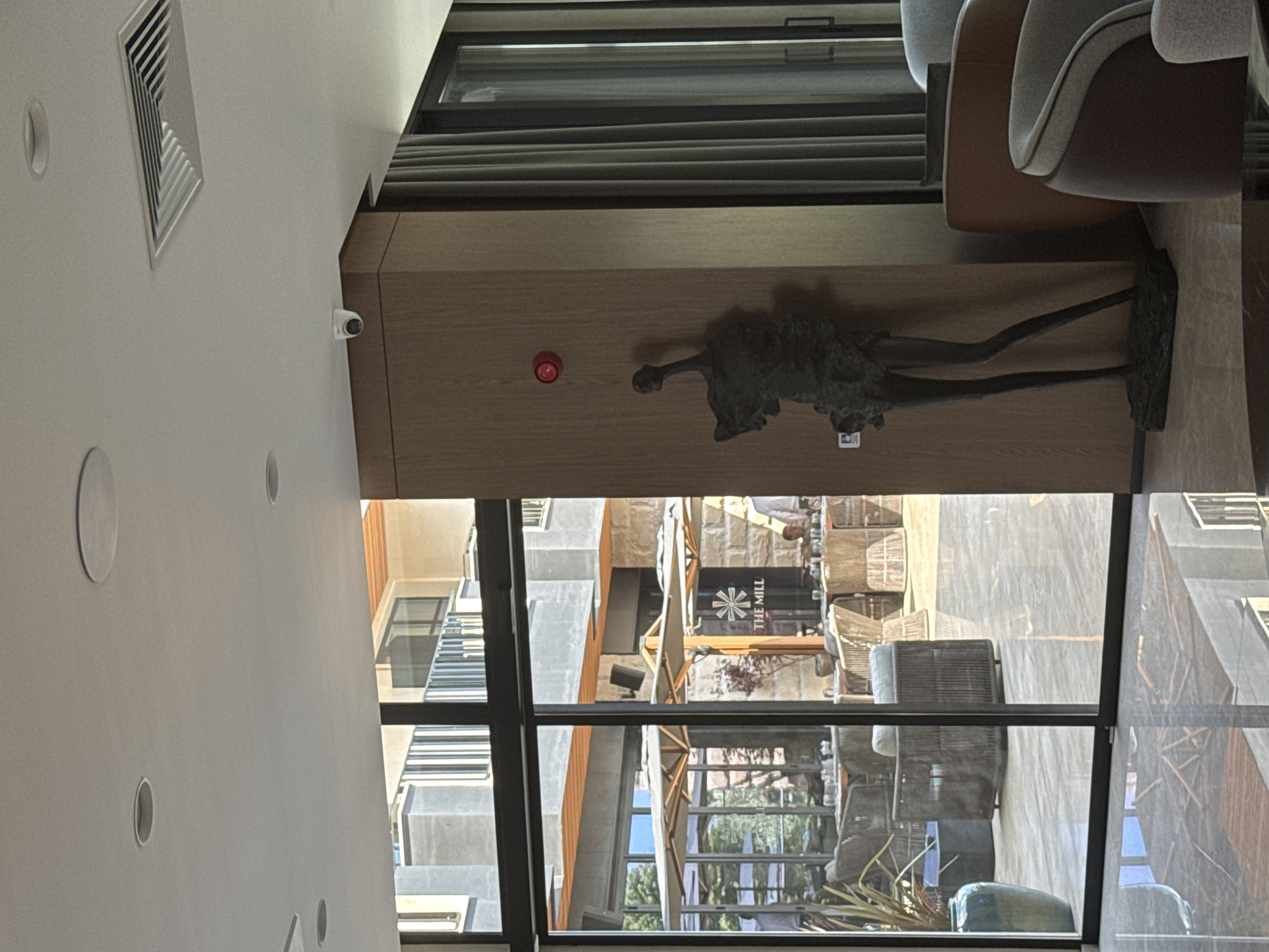 The image depicts the interior of a modern establishment, likely a hotel lobby or restaurant, in Nesebar, Bulgaria. To the left, large glass doors and windows offer a view of an outdoor seating area with tables, umbrellas, and a sign that reads "THE MILL." Inside, a dark, abstract sculpture of a human-like figure stands on a dark base against a wooden-paneled wall. A red circular object, possibly a fire alarm or emergency button, is mounted on the wall above the sculpture. Further to the right, draped grey curtains are visible alongside a glass door. The ceiling features recessed lighting and an air vent. The flooring is highly reflective, mirroring the surrounding environment. Furniture, including chairs and a large vase, is present within the interior.