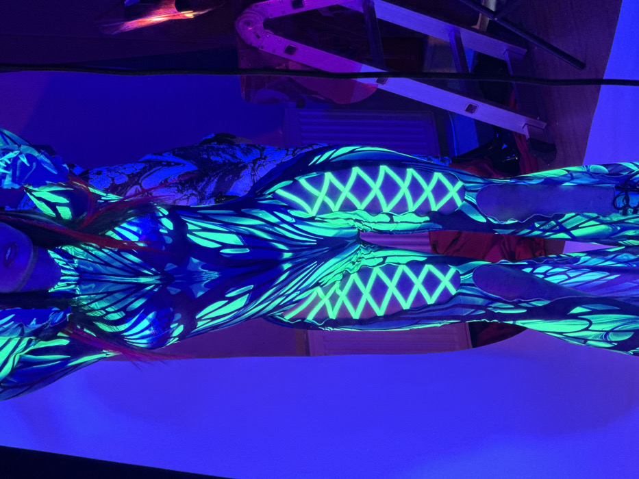 A person is positioned centrally, illuminated by ultraviolet light, causing a full-body suit to fluoresce in neon green and blue. The suit features intricate patterns, including swirling motifs on the torso, criss-cross lacing designs on the upper thighs, and flared bottoms on the lower legs. The individual's lower face, including the mouth and chin, is visible, along with parts of red or orange hair glowing under the UV light. The arms are raised above the head, with hands out of frame.
The environment is an indoor setting, likely a studio or event space, characterized by dark blue ambient light. To the left, a curved white or light-colored backdrop is visible. To the right, a white wall-mounted radiator is present. A silver A-frame ladder leans against a wall further to the right. A dark, thin vertical cable or wire hangs in the mid-ground on the right. Another patterned object or fabric, also glowing under UV light, is partially visible behind the subject's right arm. The specific location within Sofia, Bulgaria, is not discernible from the provided image.