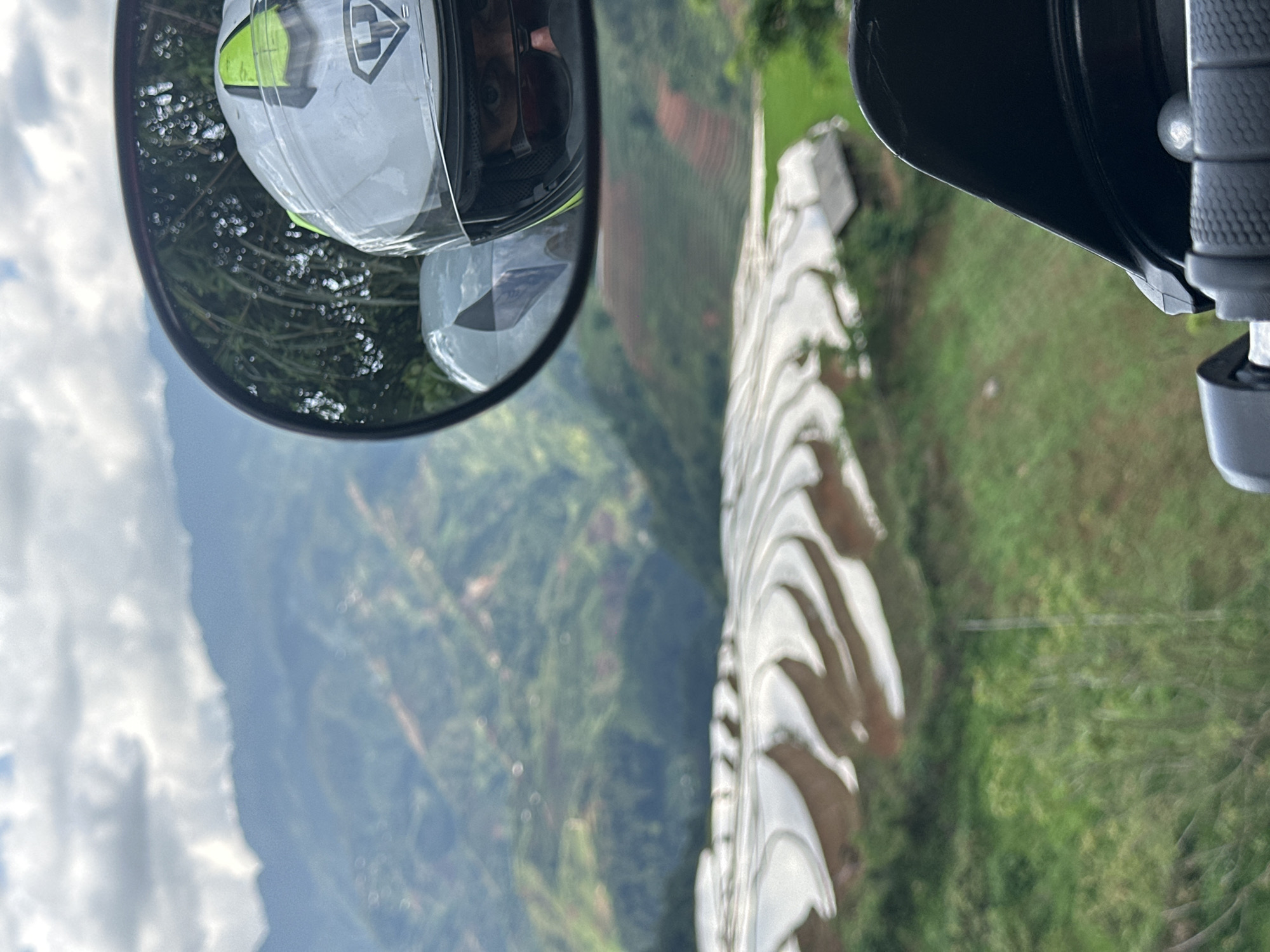 The image, taken from the perspective of a motorcycle rider, captures a scenic mountainous landscape in Lao Chai, Vietnam. It is currently displayed vertically, though the content suggests it was originally captured in landscape orientation from a moving vehicle.
In the upper left quadrant of the image, a circular rearview mirror prominently displays the reflection of the rider. The rider is wearing a white motorcycle helmet with bright green/yellow accents and a dark visor mechanism. Part of the rider's face, specifically their eyes and nose, is visible through the helmet's opening, along with what appears to be part of a headset or ear protection worn inside. The helmet also features a visible logo, a stylized shield or cross design.
To the right, partially visible, are elements of the motorcycle itself, including a black handlebar grip and a portion of the fairing or windshield.
The backdrop to this scene is a sprawling, lush green mountainous region. Valleys and distant slopes are covered in dense vegetation, with some reddish-brown areas possibly indicating exposed soil or terraced fields. The sky above is cloudy, suggesting an overcast day.
In the foreground, extending across the lower portion of the image and curving upwards, is a striking white, textured structure. It has a distinct wavy or ridged pattern, possibly a decorative wall, barrier, or architectural element, whose exact purpose isn't clear but adds a dynamic visual to the scene. The overall impression is one of travel or exploration through a beautiful, rugged environment.