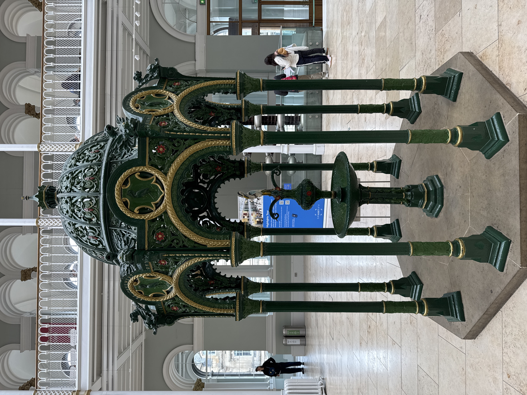 This is a medium-shot, eye-level, full-color photograph of a tall, ornate, dark green and gold fountain in the Great Hall of the National Museum of Scotland, located in the city of Edinburgh, United Kingdom. The fountain is centrally located and occupies the foreground of the image. It features a domed canopy adorned with intricate metalwork and gold accents, supported by several slender, fluted columns. Decorative circular panels depicting wildlife are integrated into the structure. A smaller basin with a bird-like sculpture sits at the base of the fountain.
The background reveals the architecture of the Great Hall, characterized by a multi-level interior with pale tiled floors, light grey walls, and large arched windows. A white balustrade with gold trim runs along an upper level, where several indistinct figures can be seen. In the middle ground, a blue sign with text and a QR code is placed to the right of the fountain, indicating a "Highlights Tour." Two silver trash receptacles are visible in the left mid-ground.
In the far left, a person wearing dark clothing and a cap stands facing away from the camera. In the middle ground, slightly to the right of the fountain, a group of three people, two women and one man, are looking at a piece of paper. The woman on the left is wearing a white top and dark pants, while the woman on the right has a yellow backpack. The man is wearing a dark top and light-colored pants. Further to the right, in the background, two people are visible in front of a large glass door, one wearing a white top and the other a darker top. The overall environment suggests an indoor public space, likely a museum or gallery, with a bright, diffused lighting.