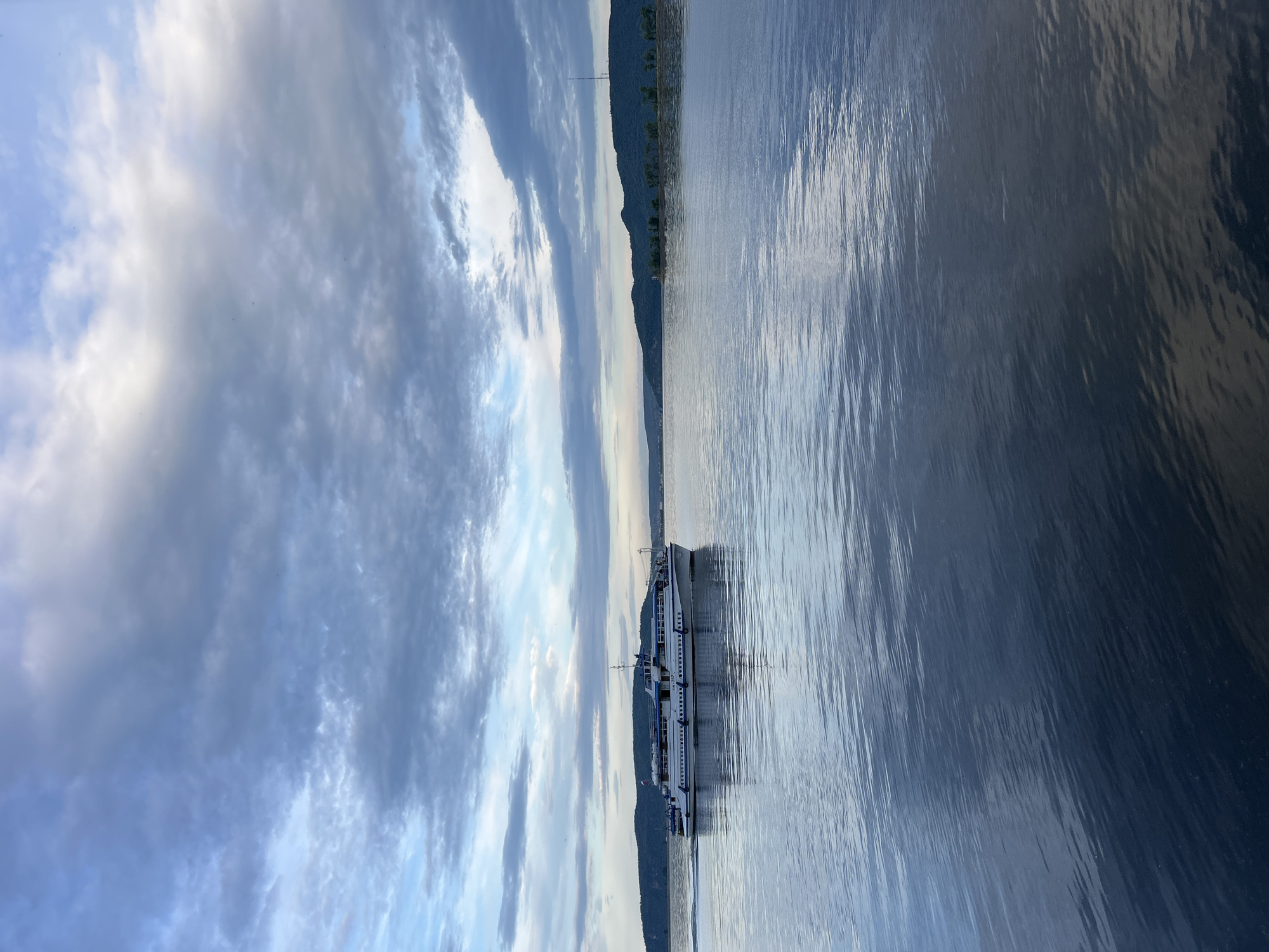 The image depicts a ferryboat navigating a body of water, with a silhouetted shoreline and a dramatic sky. The ferry, a long, multi-decked vessel with a white and blue color scheme, is positioned to the left of the frame, cutting through the water. Its reflection is visible on the rippled surface. The water itself appears calm, with a metallic sheen that reflects the sky.
To the left of the water, a dark, undulating shoreline is visible, suggesting a natural landscape with trees or vegetation. A tall transmission tower rises from the land, breaking the silhouette. The sky is filled with a mix of dark, dramatic clouds and lighter, brighter patches, hinting at either sunrise or sunset, or a partly cloudy day. The overall mood is serene and somewhat dramatic, with the vastness of the sky and water dominating the scene.
There is no visible text in the image. The time of day appears to be either early morning or late evening, given the quality of light and the cloud formations. The location is Tolyatti, Russia, which implies this scene could be on the Volga River or one of its reservoirs. The activity is simply the ferry in transit across the water.