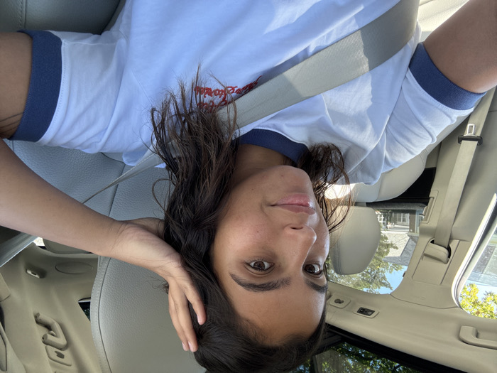 A young adult female is depicted in a medium close-up, seated in the front passenger seat of a light-colored vehicle, secured by a light gray seatbelt. She has medium-length dark hair, styled to the side, and is looking directly at the camera with a subtle smile. Her right hand is raised, with fingers touching the hair above her ear. She is wearing a white short-sleeved ringer t-shirt with blue trim on the collar and sleeves. Red embroidery, partially obscured, is visible on the front of her shirt. The car's interior features light-colored upholstery, likely leather, and a panoramic sunroof is visible overhead, allowing bright sunlight into the cabin. Through the vehicle's windows, a residential scene is visible under a clear blue sky, featuring a large house with light siding and a dark roof, surrounded by green foliage, consistent with a suburban environment in Princeton, United States.