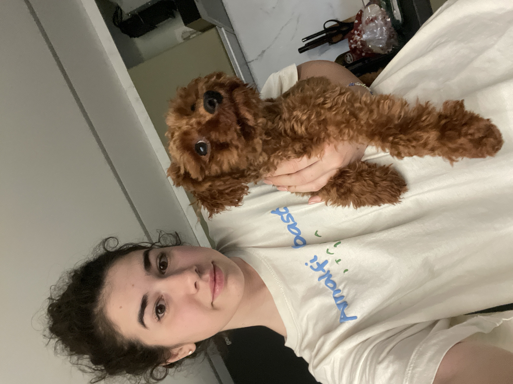 A person with curly dark hair is holding a small, brown, curly-haired dog. The person is wearing a white t-shirt with "Amalfi Coast" printed in blue. The setting appears to be indoors, potentially a residence. There is no information to suggest the location is in Sofia, Bulgaria.