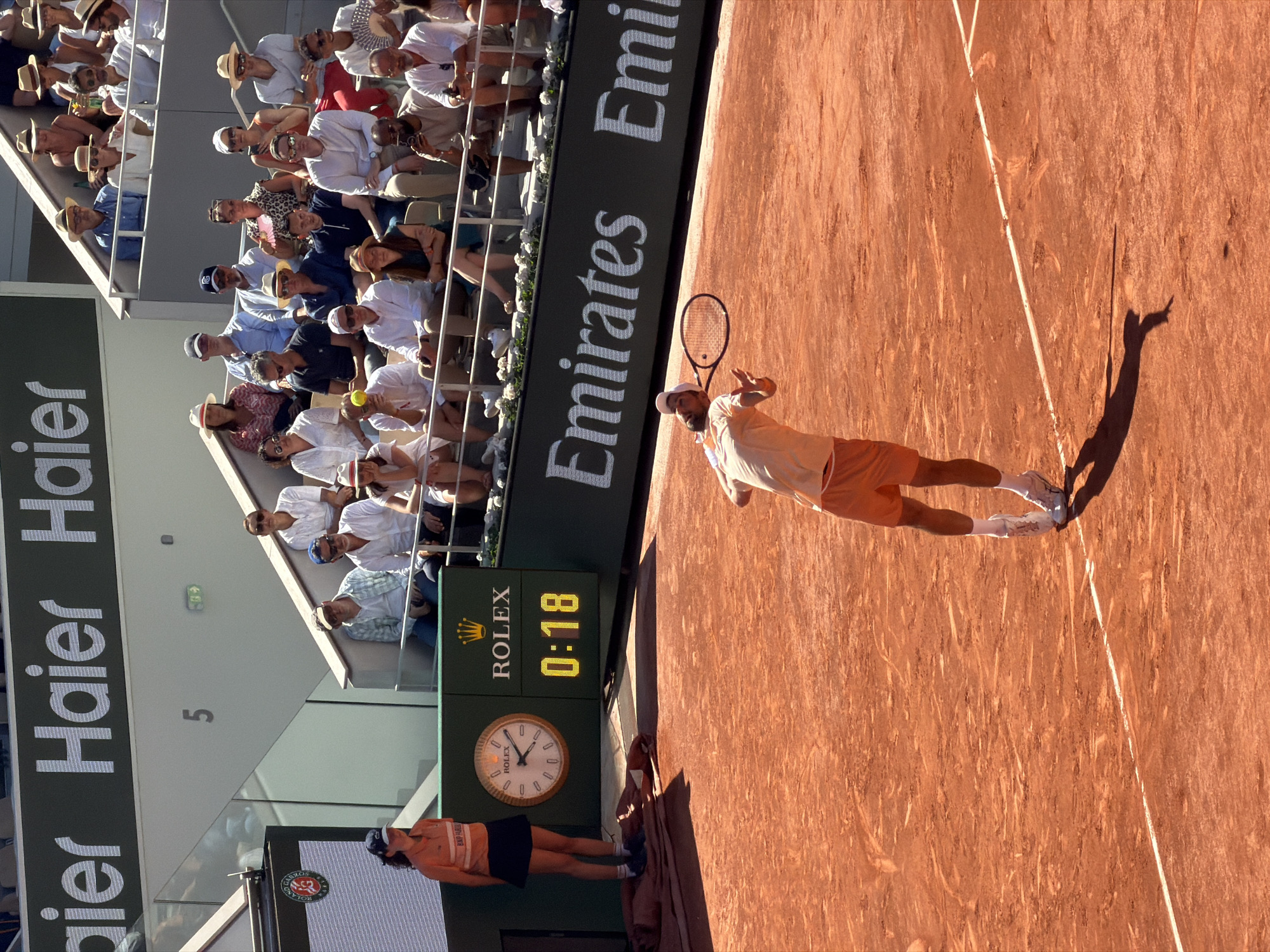 The image captures a tennis match in progress at Roland Garros in Paris, France. A male tennis player, wearing an orange shirt and shorts, white socks, and shoes, is in the midst of serving. His shadow is cast onto the red clay court. A line judge, dressed in a navy skirt and an orange top with white shoes, stands near the court. A large advertising sign is visible along the edge of the court: "Emirates." Behind this sign is a seating area filled with spectators. Above the line judge is a screen showing a clock and displaying the time as 0:18, with the word "ROLEX" visible. In the background, there's a scoreboard with "Haier" printed vertically. Number 5 is written on the wall of the stands.