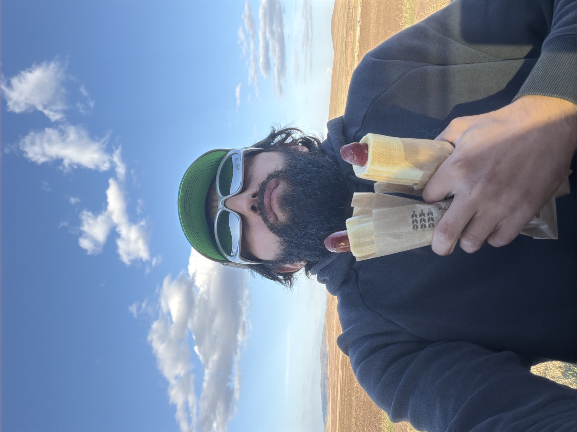 A man with a beard and sunglasses is holding two hot dogs in brown paper wrappers. He is wearing a green hat and a dark sweatshirt. The background shows a blue sky with clouds and a brown field. The location is Первомайское, Russia.