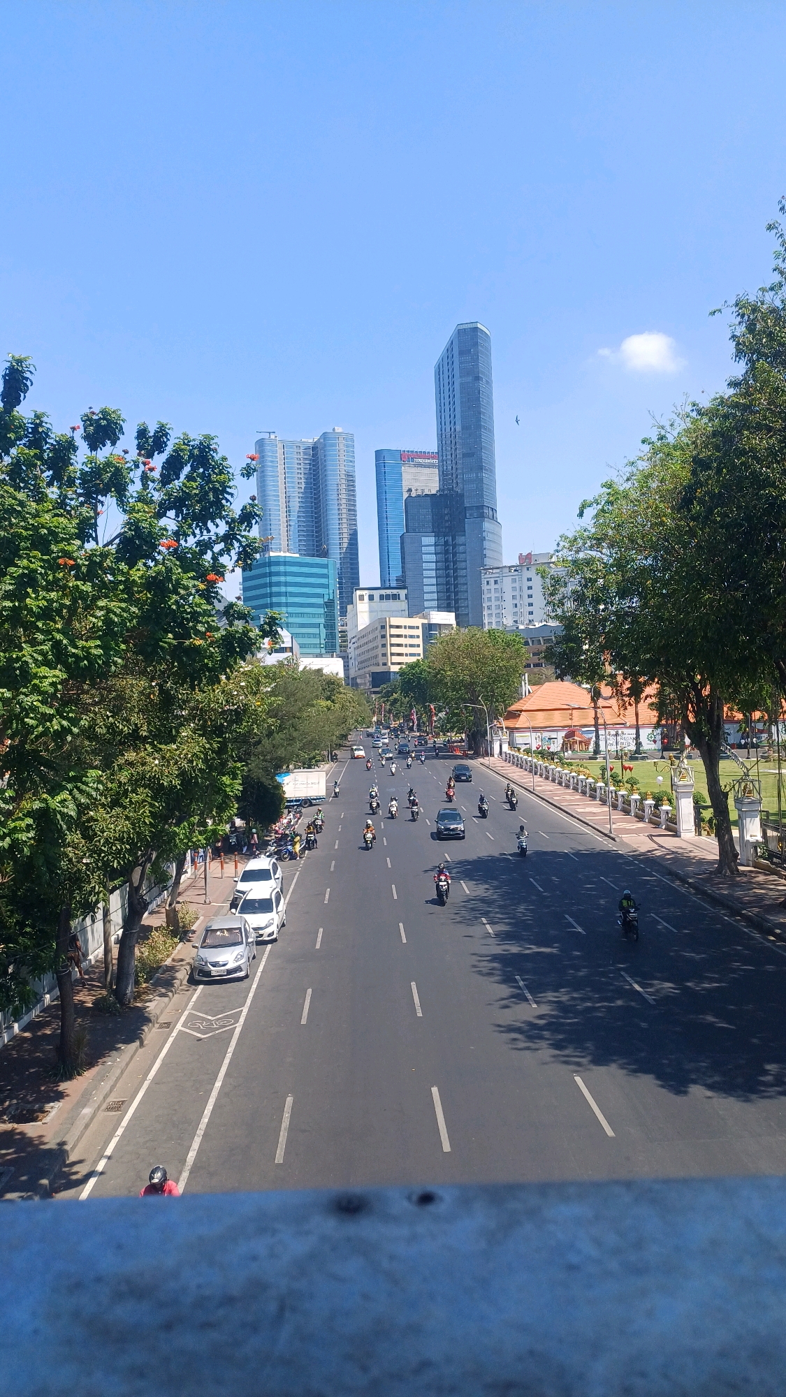 This is an eye-level, medium-shot, outdoor photograph taken on a clear, sunny day, looking down a multi-lane road in Surabaya, Indonesia. The perspective is from a bridge or elevated walkway. The road is busy with traffic, primarily motorcycles and a few cars. The architecture suggests a modern city with several tall, glass-fronted buildings visible in the background. On the right side of the road, there is a park-like area with a traditional-style building with an orange roof, surrounded by a white fence and manicured grass. Lush green trees line the left side of the road and also feature on the right, partially obscuring the view of the buildings. The overall impression is of a vibrant, bustling urban environment in Surabaya.