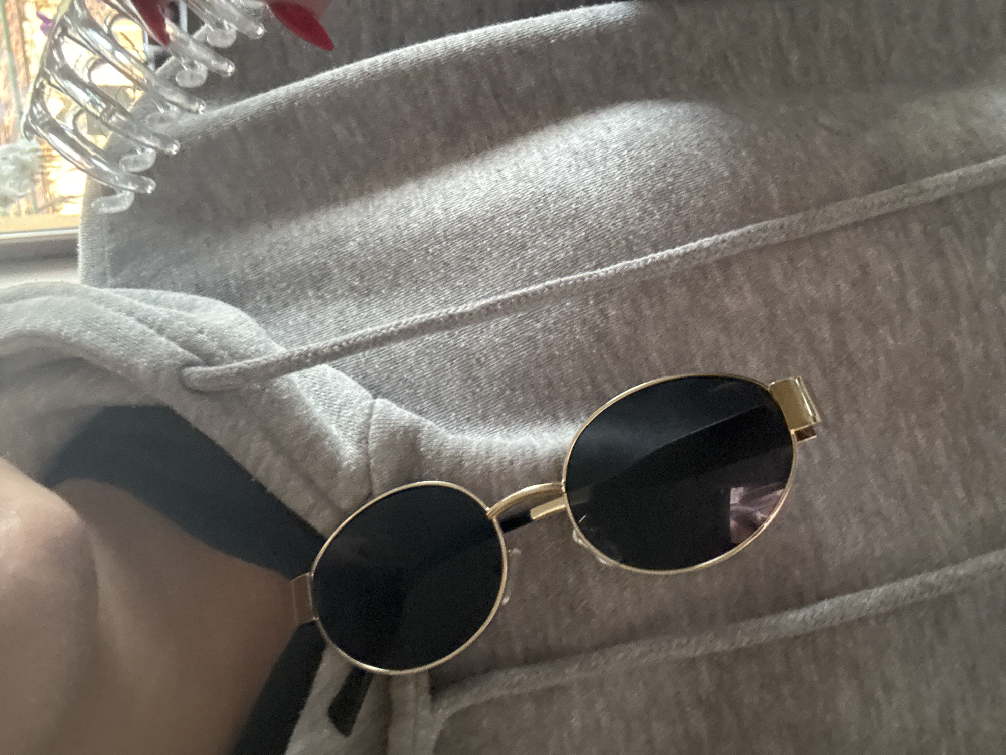 The image shows a pair of gold-framed, round sunglasses resting on a grey hooded sweatshirt. Part of a clear plastic hair clip is visible in the upper left corner. A sliver of skin is visible to the left. The location is not identifiable as Lehchevo, Bulgaria.
