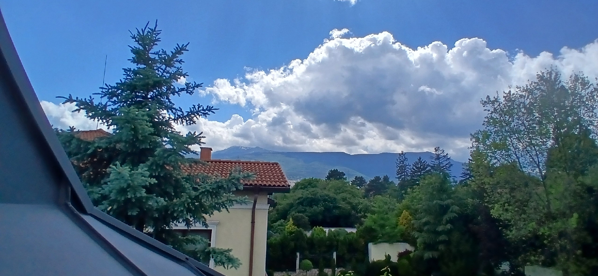 The image captures a scene from Sofia, Bulgaria, featuring a partly cloudy sky above a landscape of buildings and trees. In the foreground, a section of a dark-colored roof is visible on the left, while a green spruce tree stands nearby. A building with a red tiled roof and light-colored exterior walls with a chimney is positioned between the spruce tree and the other trees. In the midground, various trees, with different shades of green, fill the frame, some hiding small buildings. In the distance, mountains are visible, partially covered with snow under the clouds. The sky occupies the upper half of the image, filled with white and gray clouds against a blue sky.