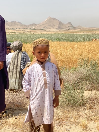 A young boy wearing a patterned white shirt and a light brown prayer cap stands in a field near Mir Ahmad Kalay, Afghanistan. Behind him, several men stand together, some wearing traditional Afghan clothing and turbans. In the background, a golden wheat field stretches towards a landscape with a large hill or small mountain under a hazy sky. Some green fields are visible in the distance. The immediate foreground shows dry, sparse vegetation.