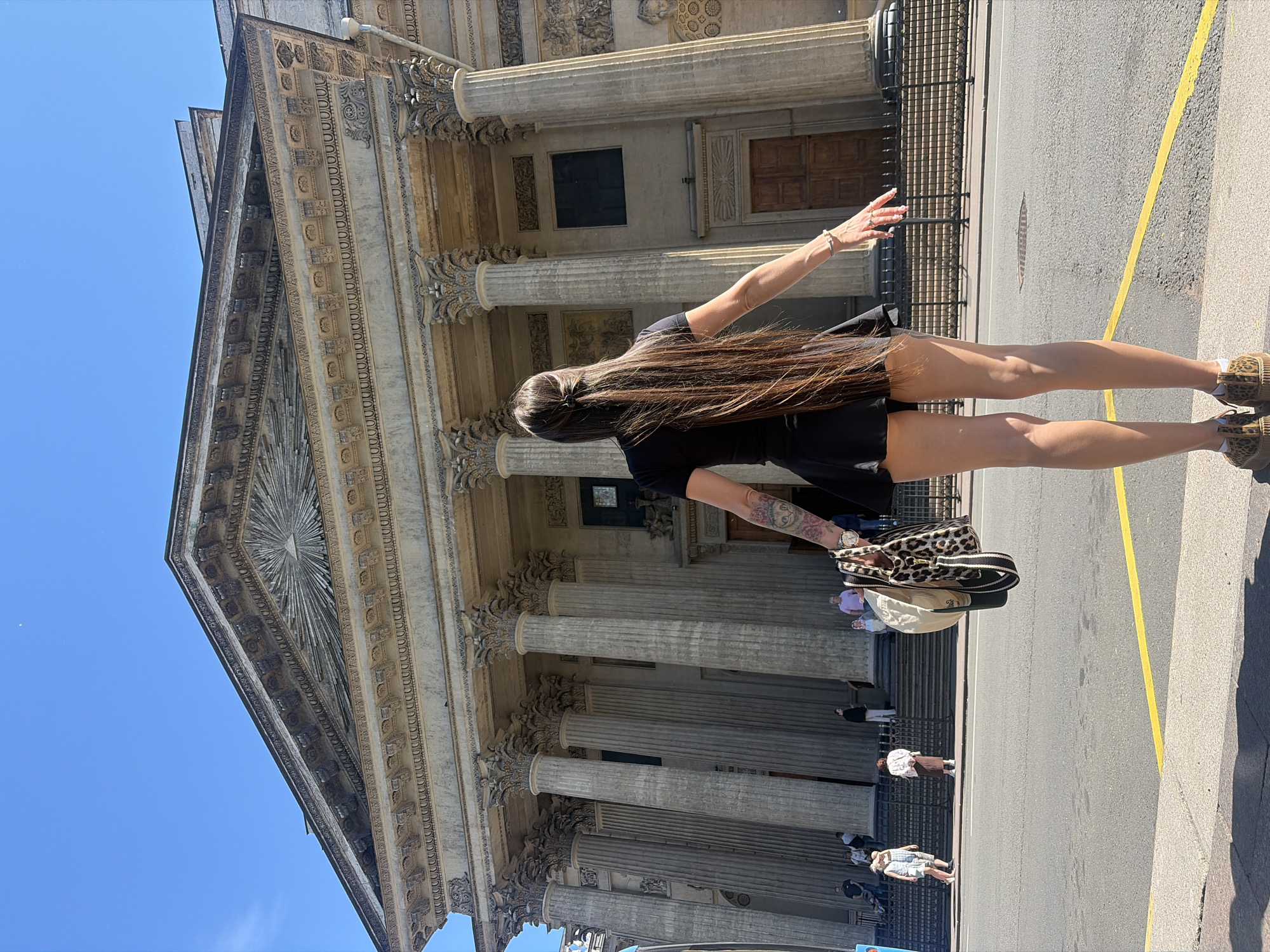The image shows a woman standing on a street, facing away from the camera, with a large neoclassical building behind her. The building features a prominent pediment with a sunburst motif, Corinthian columns, and large wooden doors. The woman is wearing a black dress, leopard-print sneakers, and is holding a beige hat and a leopard-print bag. Several other people are visible on the steps leading up to the building. The scene is set on a sunny day with a clear blue sky. The building is identifiable as the Kazan Cathedral in Saint Petersburg, Russia.